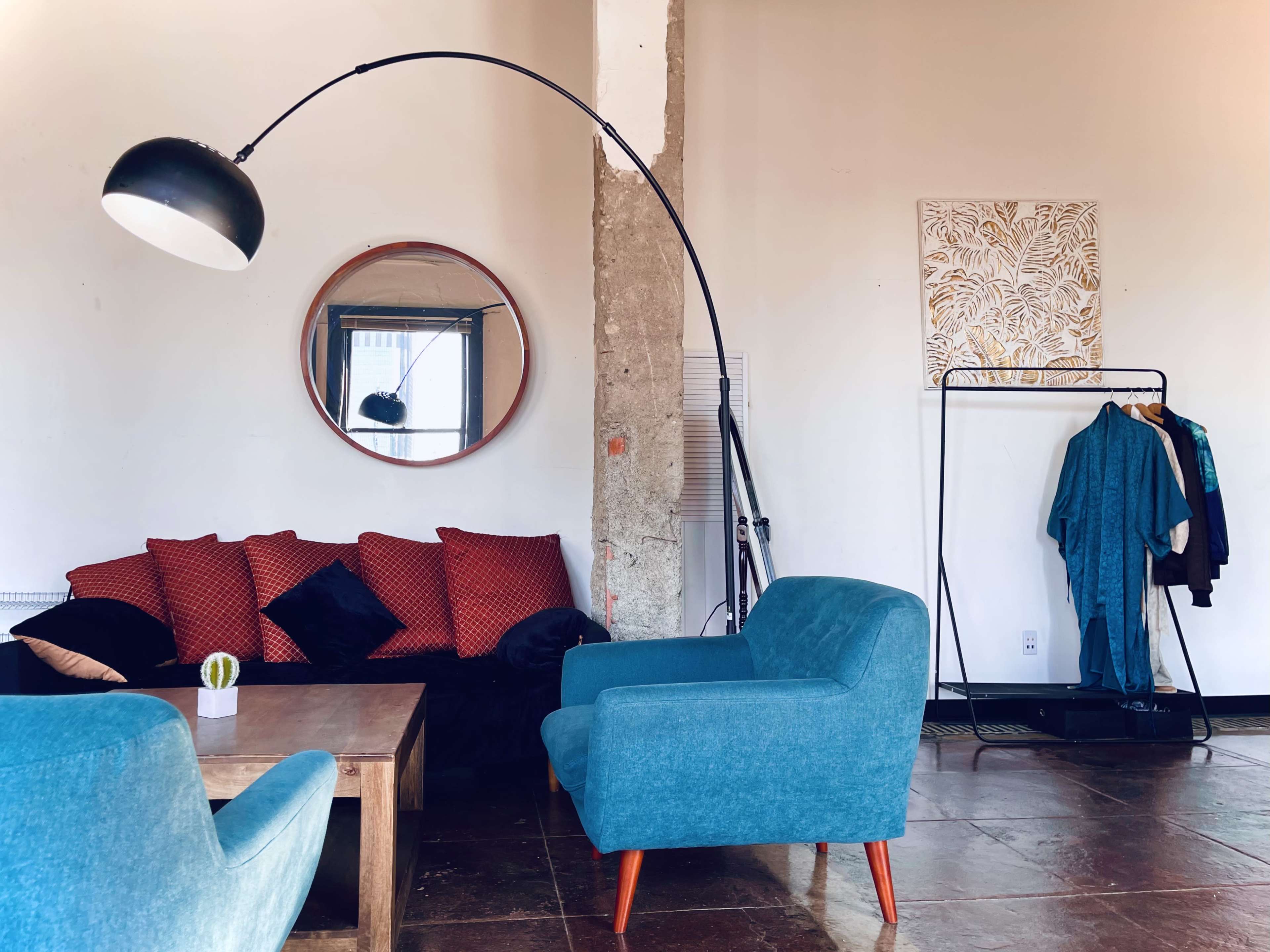 The scene features a modern living room with a circular mirror, a red and black sofa, two blue armchairs, a wooden coffee table, and a clothing rack.