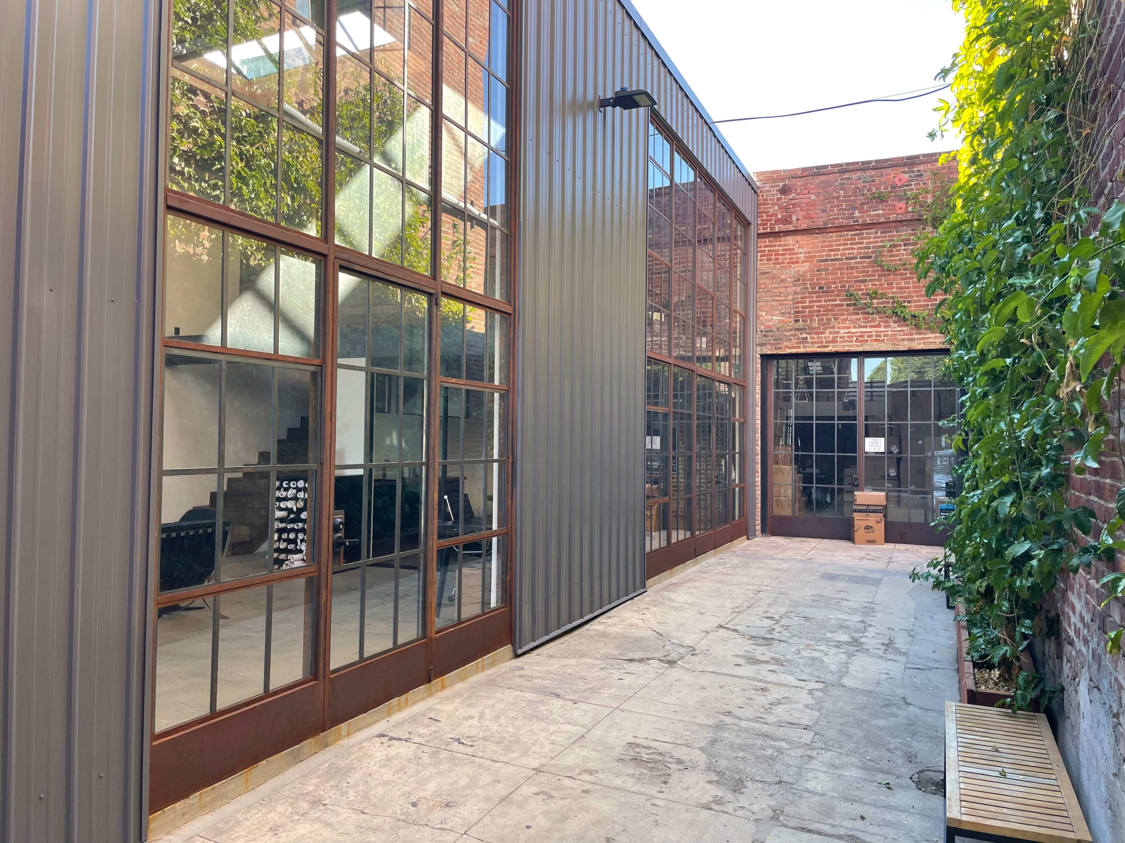 A narrow courtyard flanked by buildings with large glass windows and exposed brick walls, featuring a concrete pathway and greenery along one side.