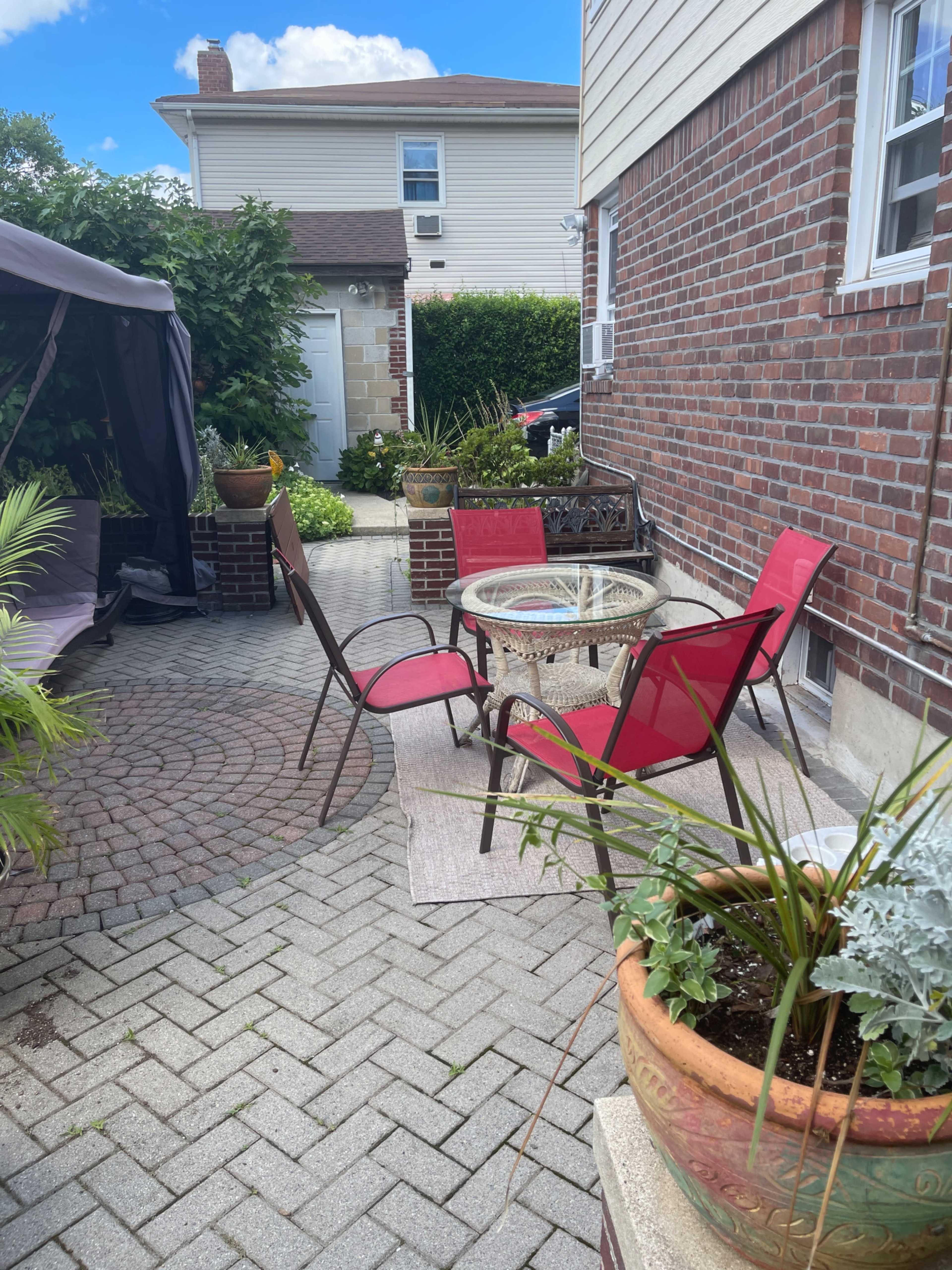 A small patio area features a round glass table surrounded by four red chairs, with brick walls and greenery in the background.