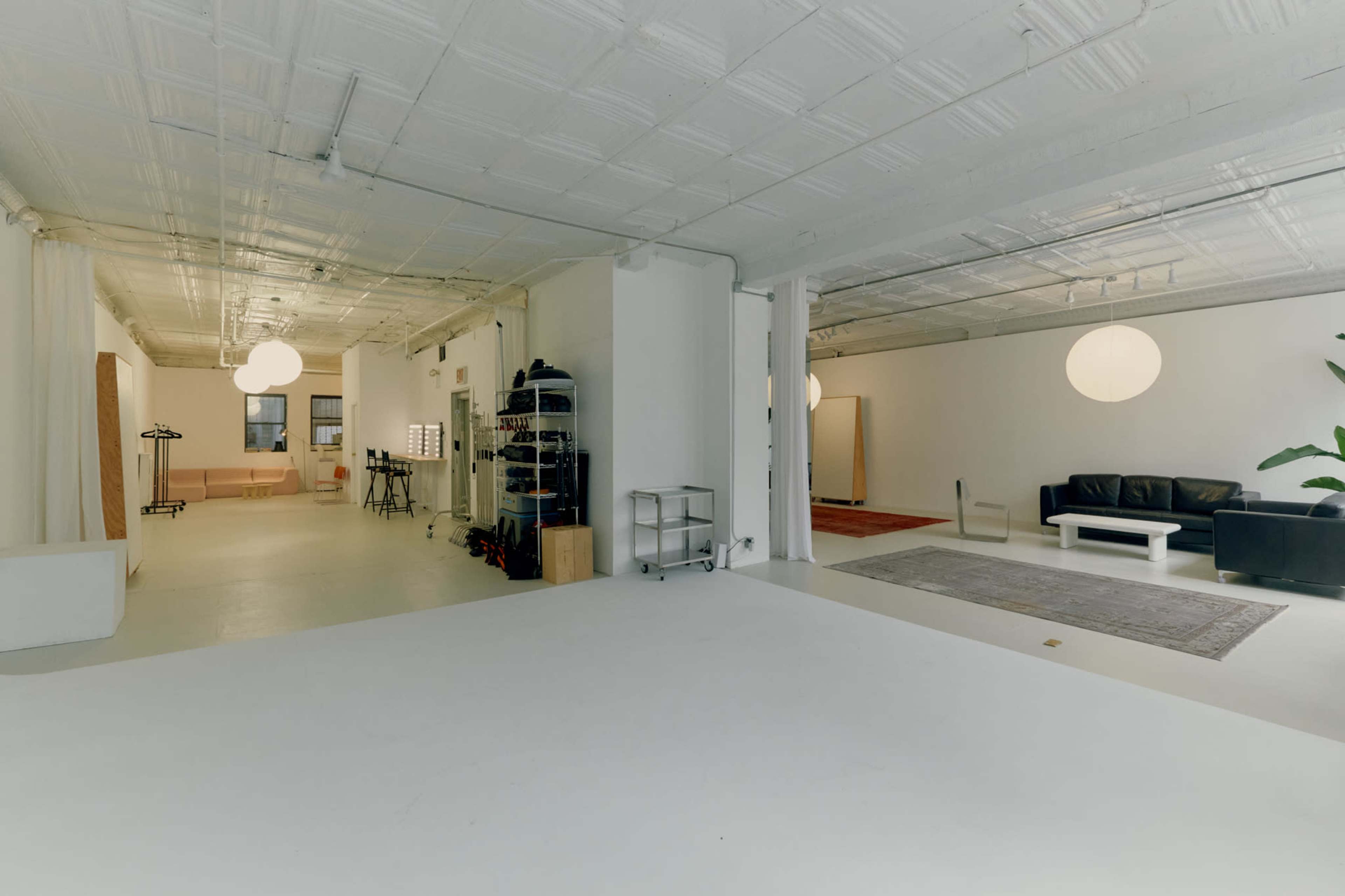 The image shows a spacious, brightly lit studio with white walls, a seating area, and a black leather couch, separated by open space and minimal decor.