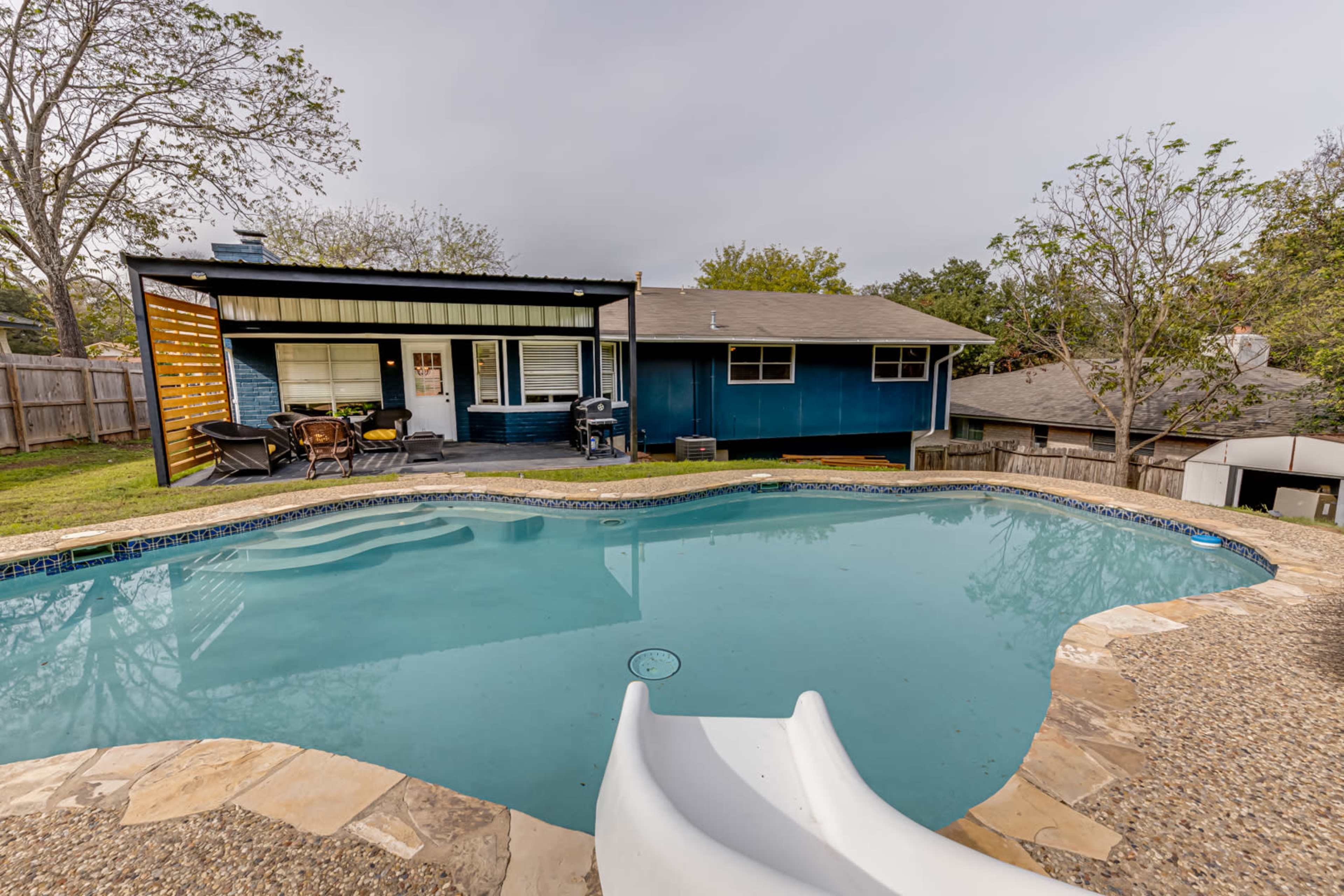 A backyard scene featuring a swimming pool with a slide, adjacent to a single-story house with a patio and yard.