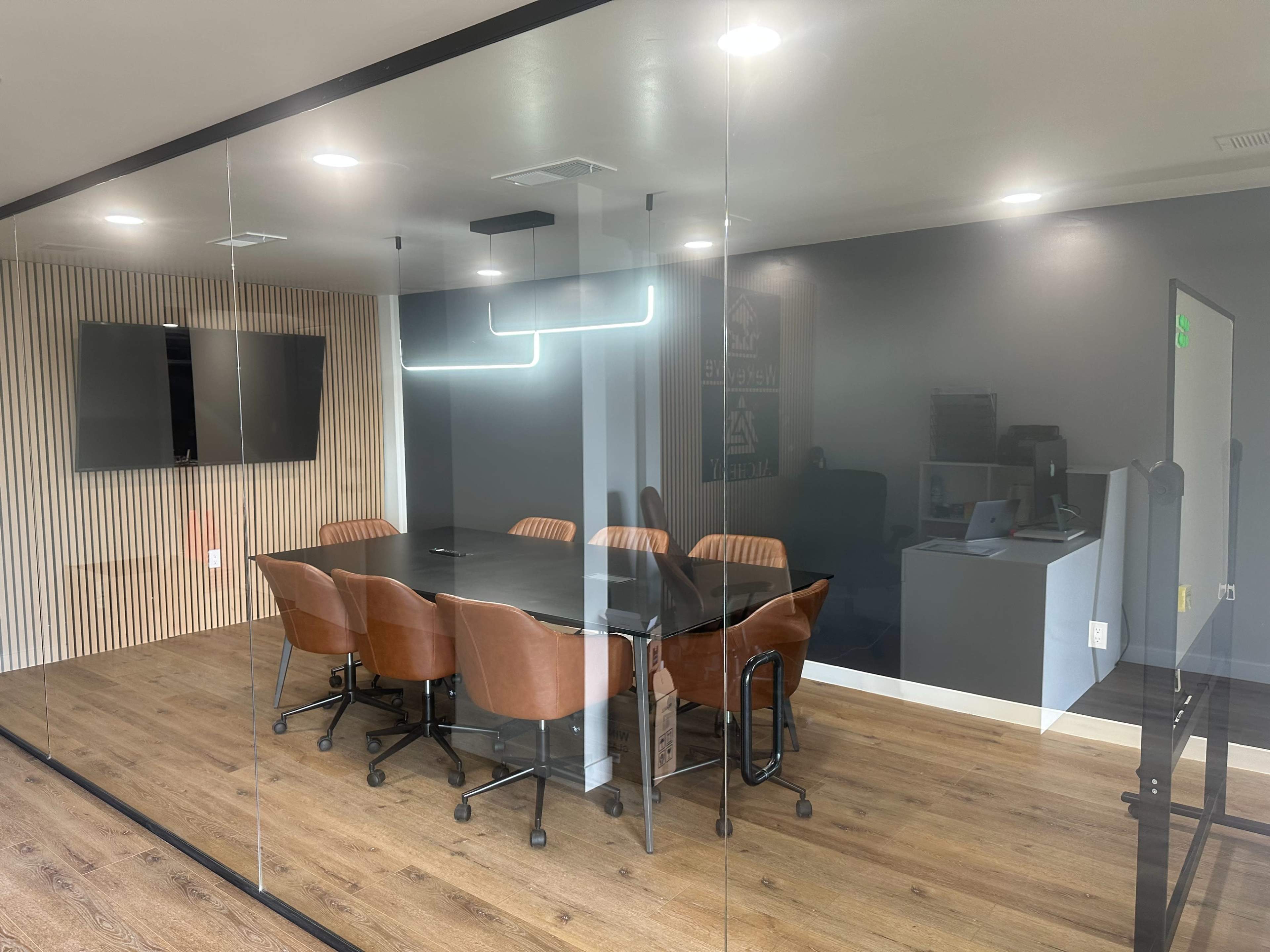 A modern conference room with a large black table surrounded by brown chairs, glass walls, and a wall-mounted TV.