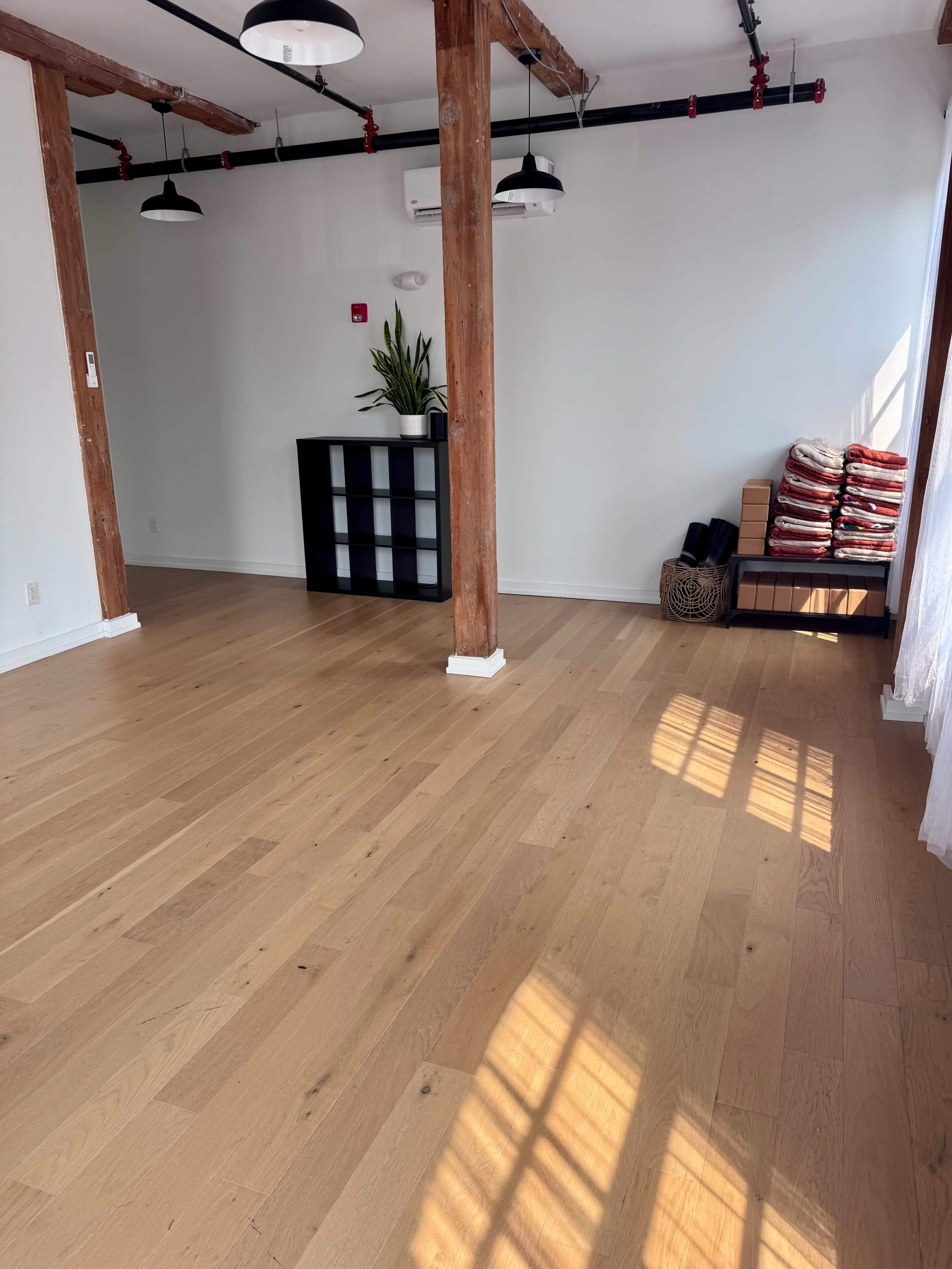 The image shows an empty room with wooden floors, exposed wooden beams, and minimal furniture, including a black shelf and neatly stacked items against the wall.