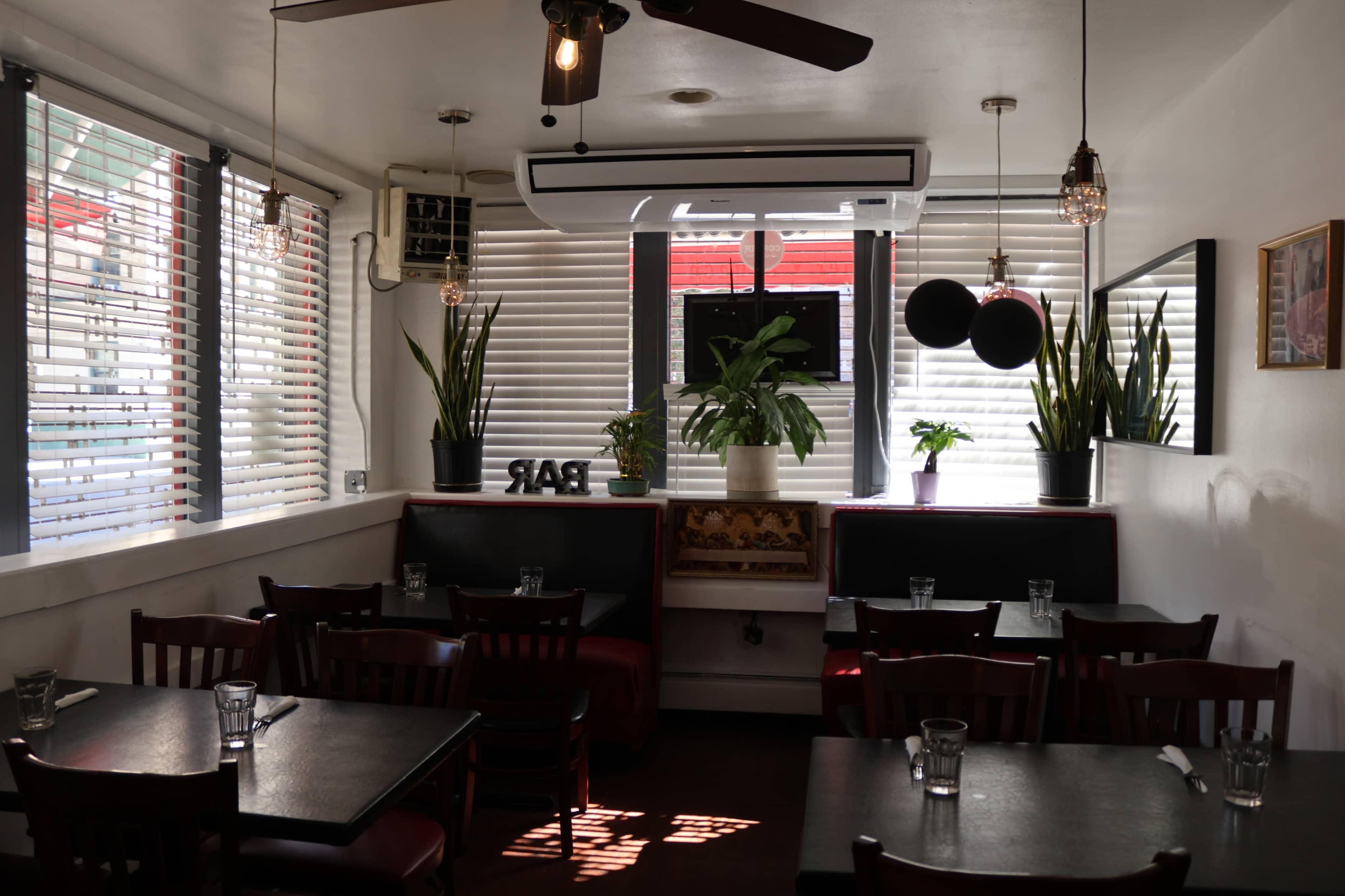 The image shows a modern restaurant interior with several tables, plants, and large windows covered by horizontal blinds.