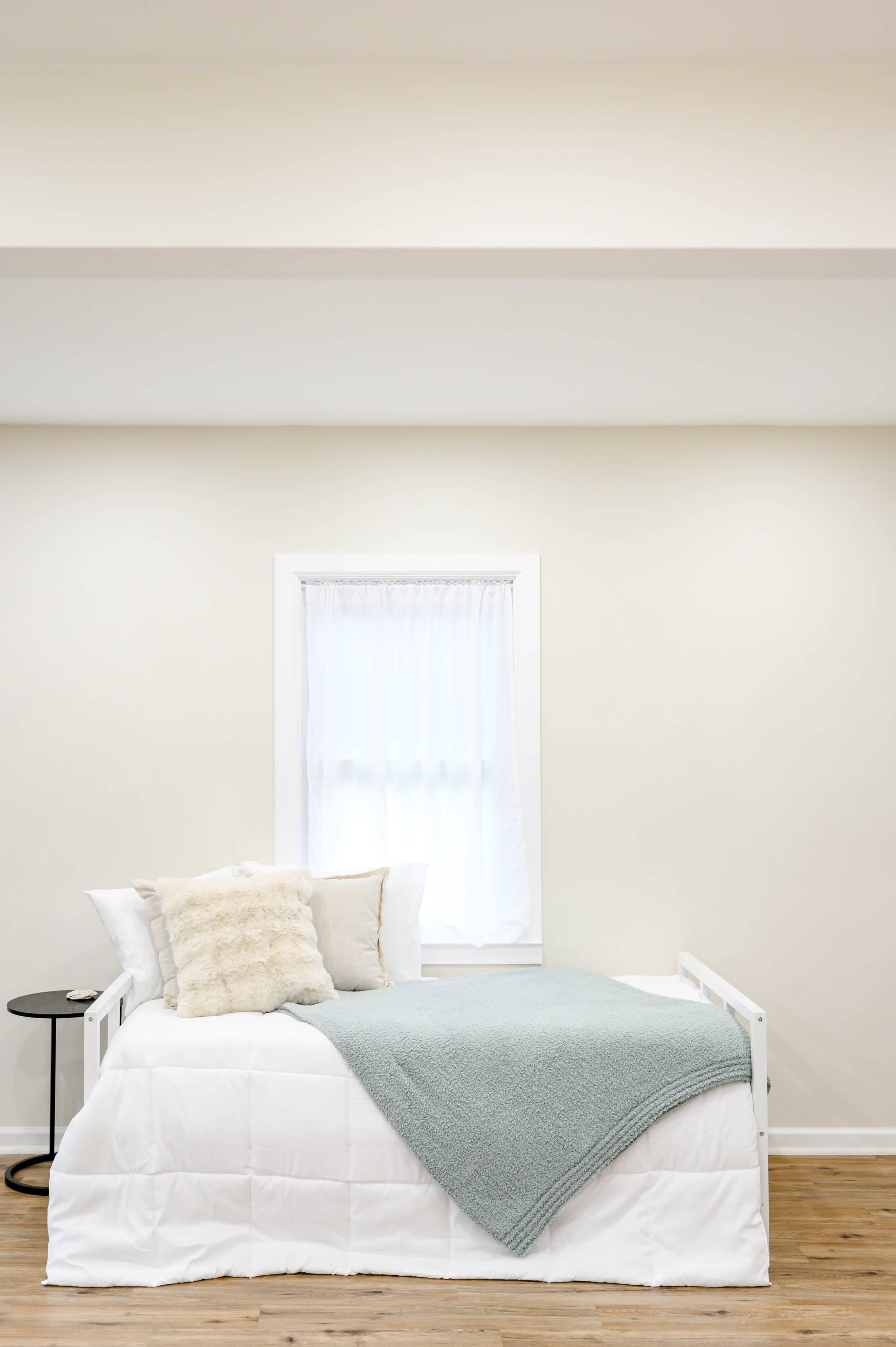 The image shows a neatly arranged bedroom featuring a bed with a light blanket and decorative pillows, a small black side table, and a window with white curtains.