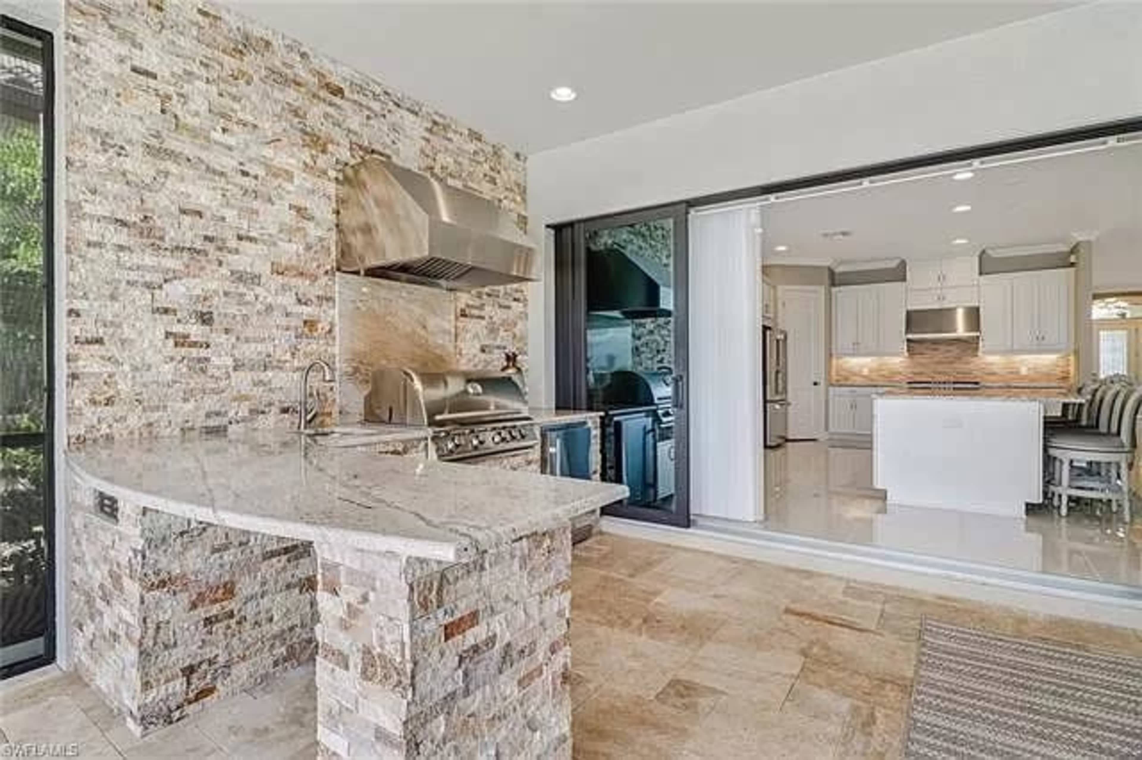 A modern outdoor kitchen with a stone grill area and an open layout connecting to a sleek indoor kitchen.