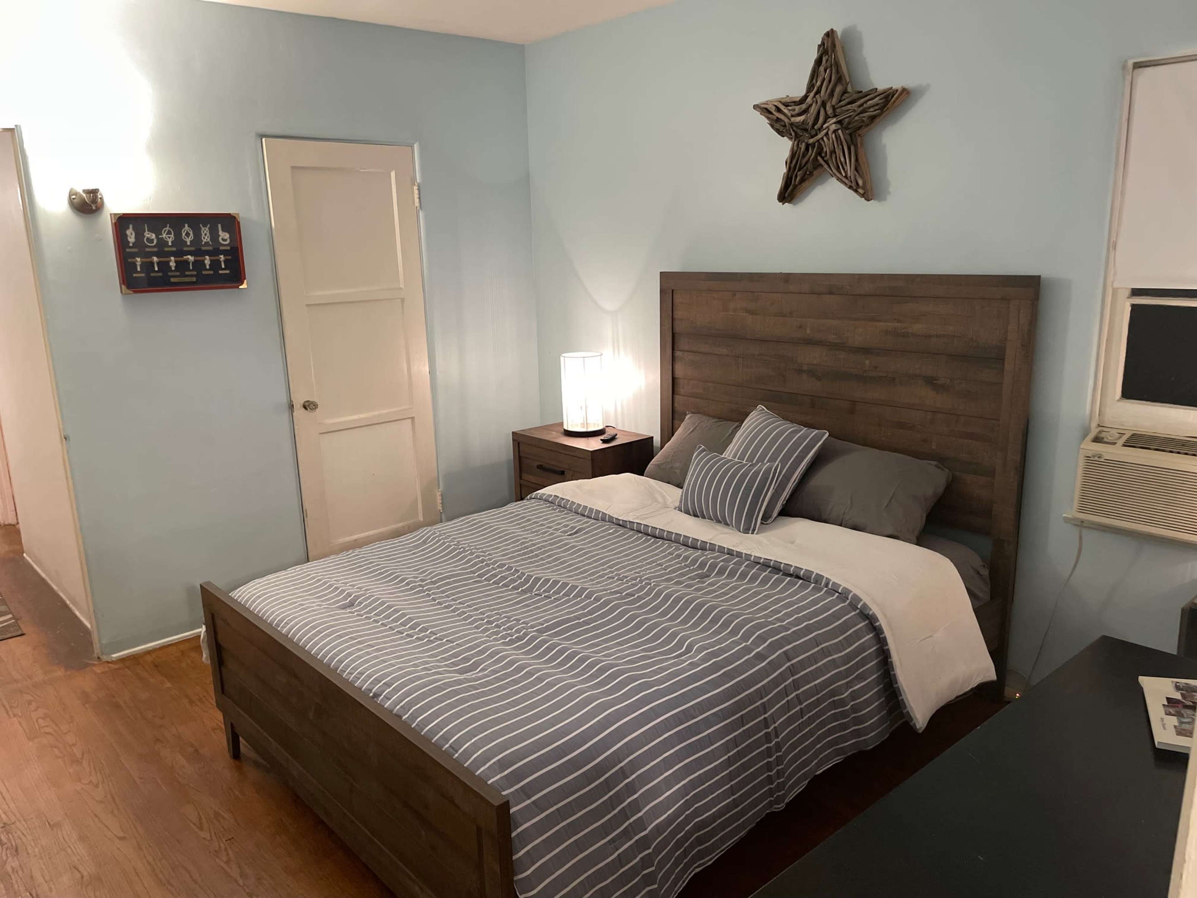 The image shows a simple bedroom featuring a wooden bed frame with striped bedding, a nightstand, and a star-shaped decor piece on the wall.