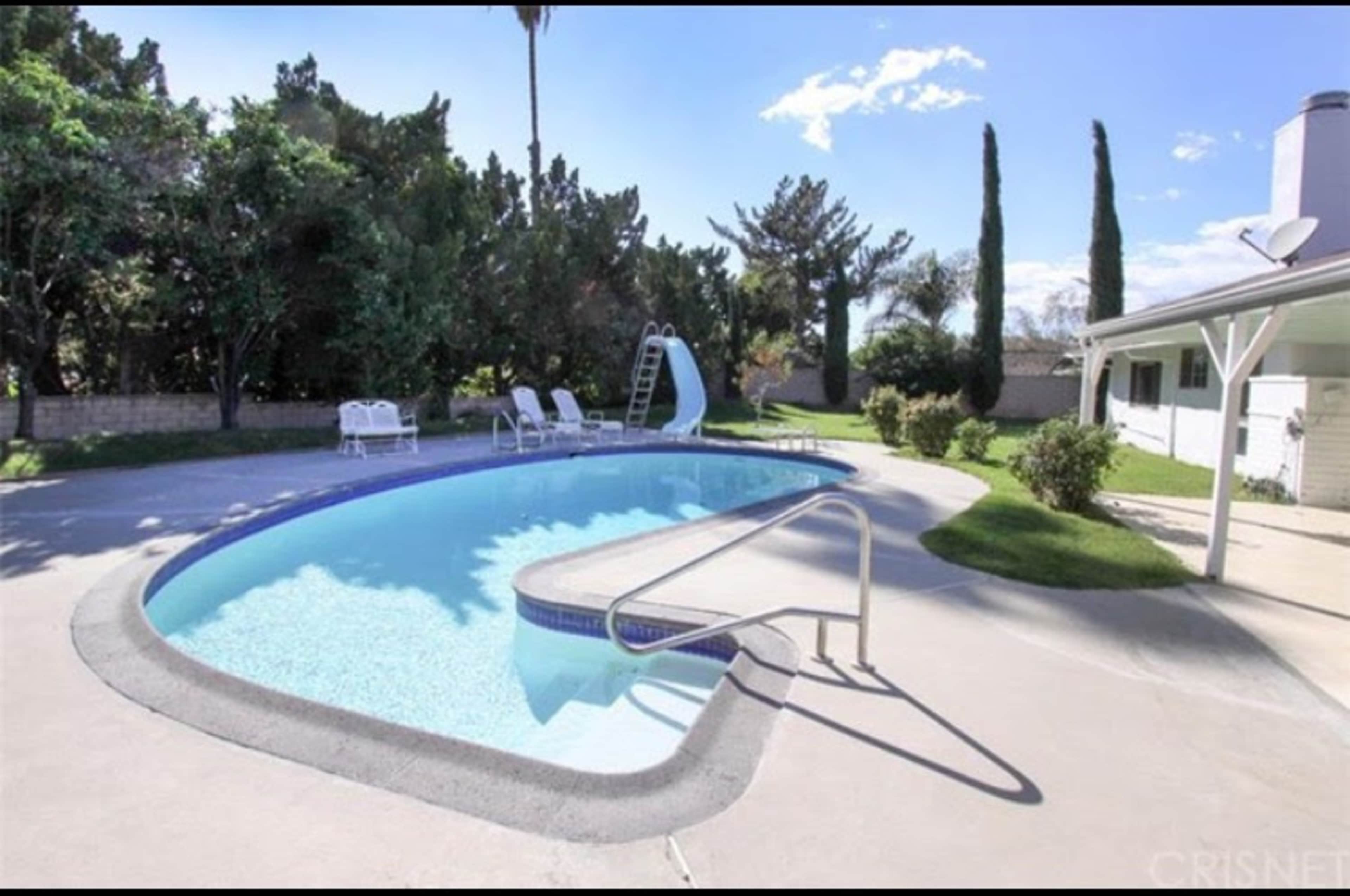 A swimming pool surrounded by green trees and lawn chairs, with a slide at one end.