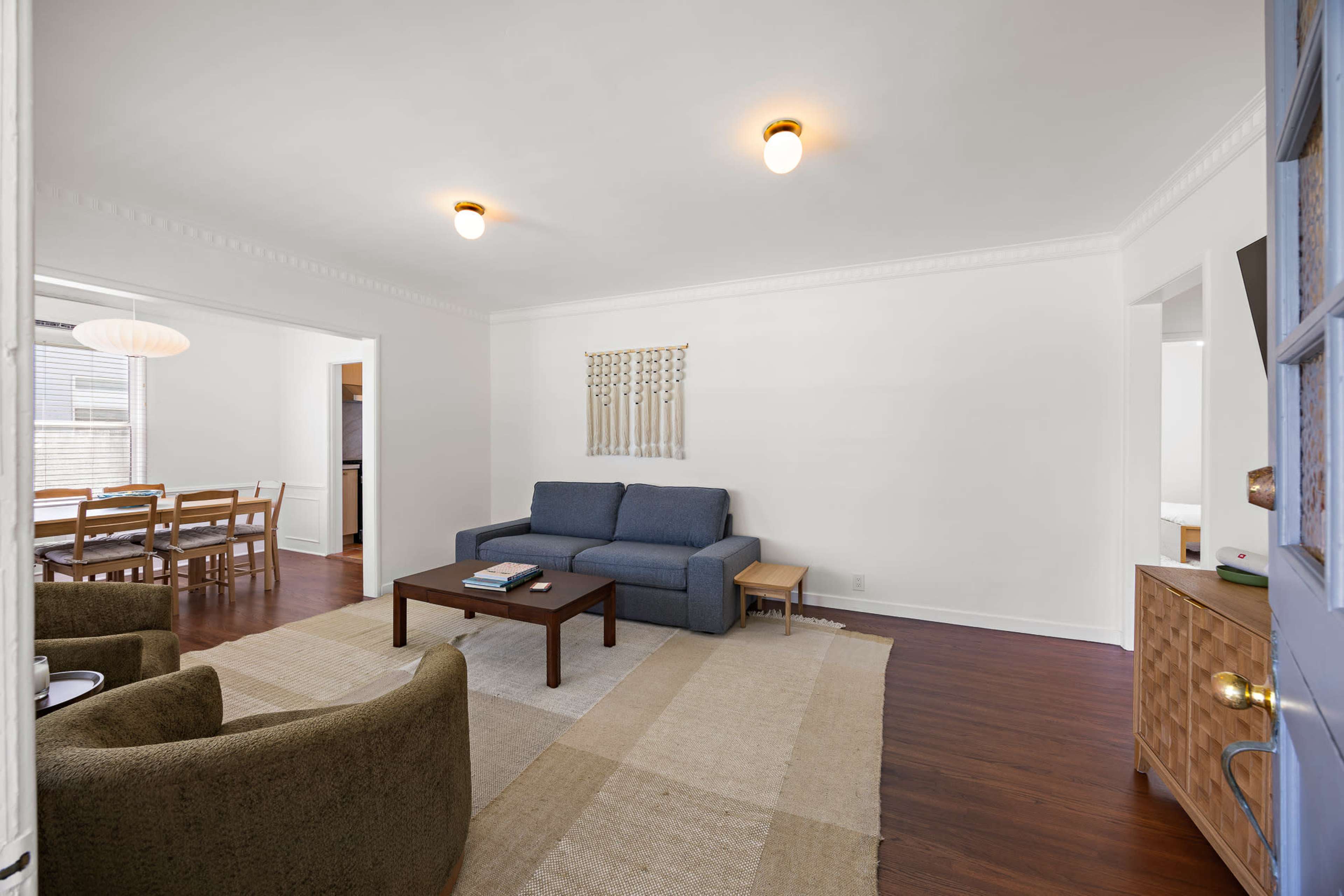 The image shows a spacious, well-lit living area featuring a blue sofa, a brown coffee table, and a seating arrangement including two armchairs, with a dining area visible in the background.