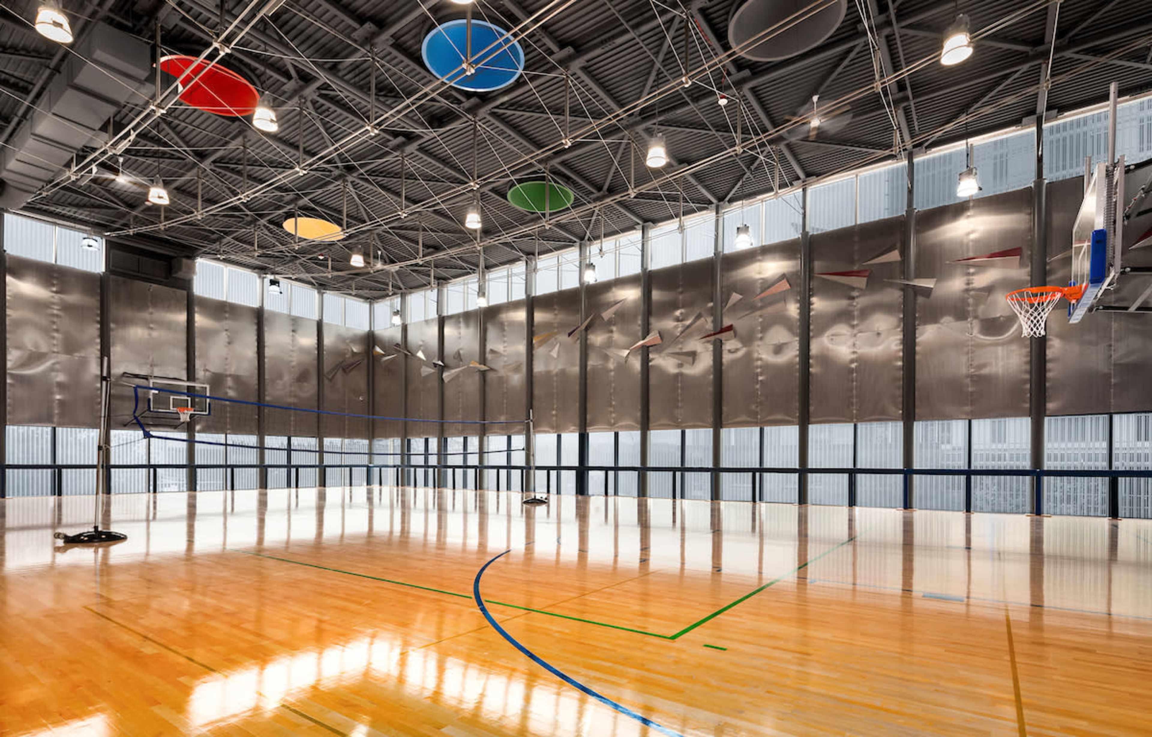 The image shows an indoor sports gym with a polished wooden floor, a basketball hoop, and colorful acoustic panels on the ceiling.