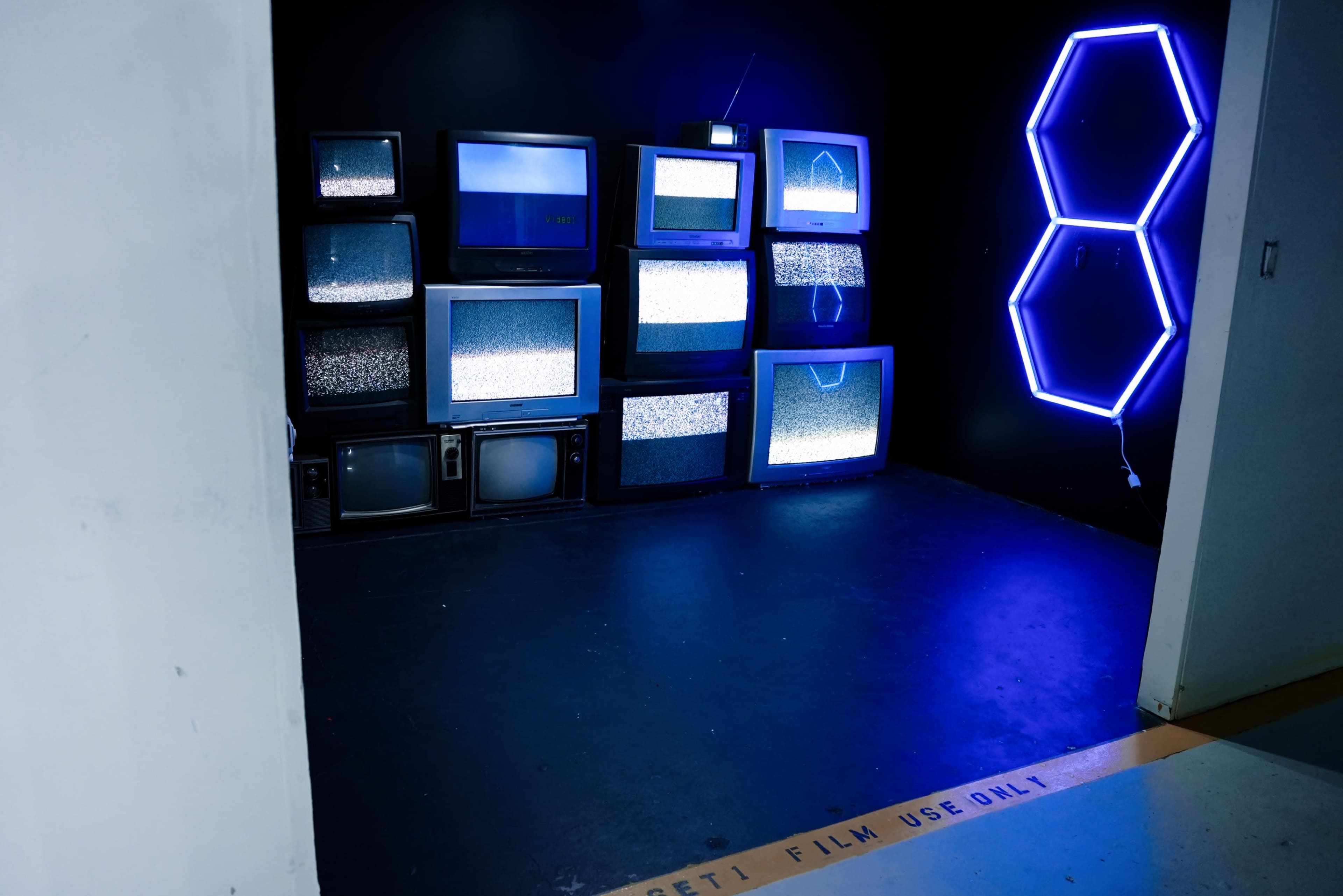 The image shows a dark room featuring multiple stacked vintage televisions displaying static on a black wall, with a neon hexagon light illuminating the space.