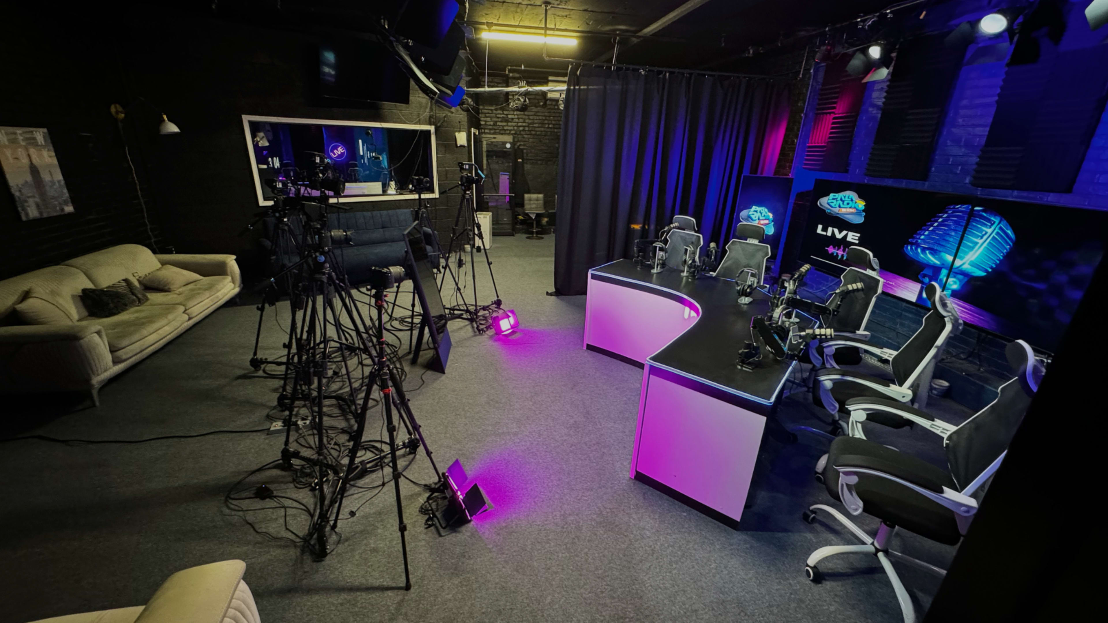 A television studio setup with multiple camera tripods, a desk with gaming chairs, and purple-lit background elements.