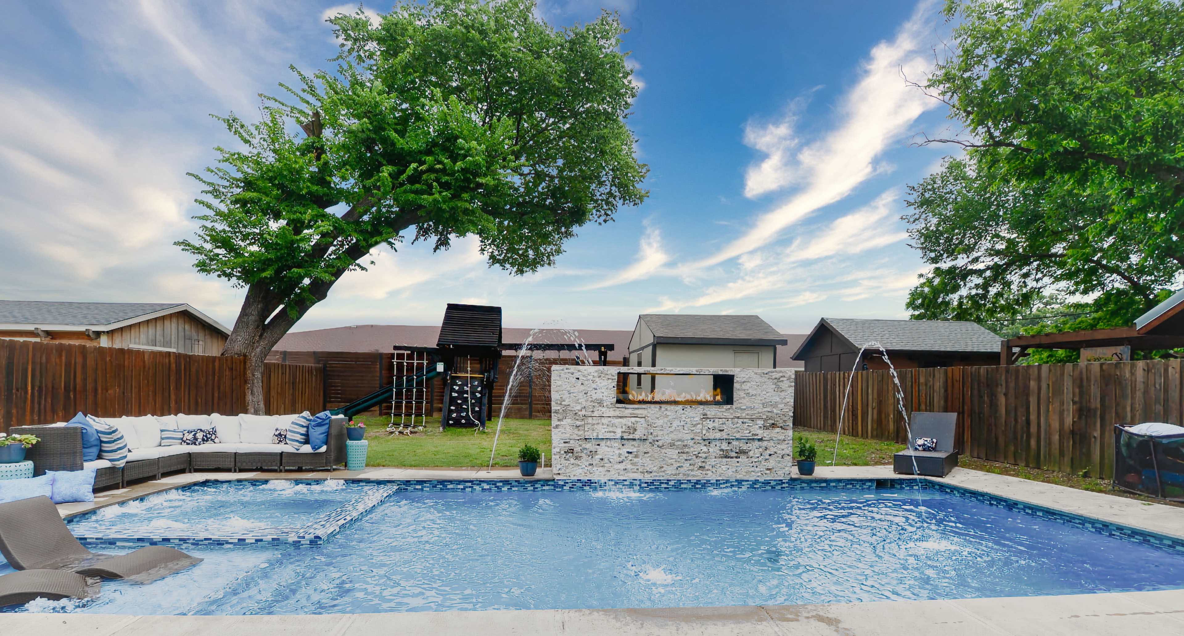 The image shows a backyard pool area with a stone wall fountain, lounge chairs, and a playset in a fenced garden under a blue sky.