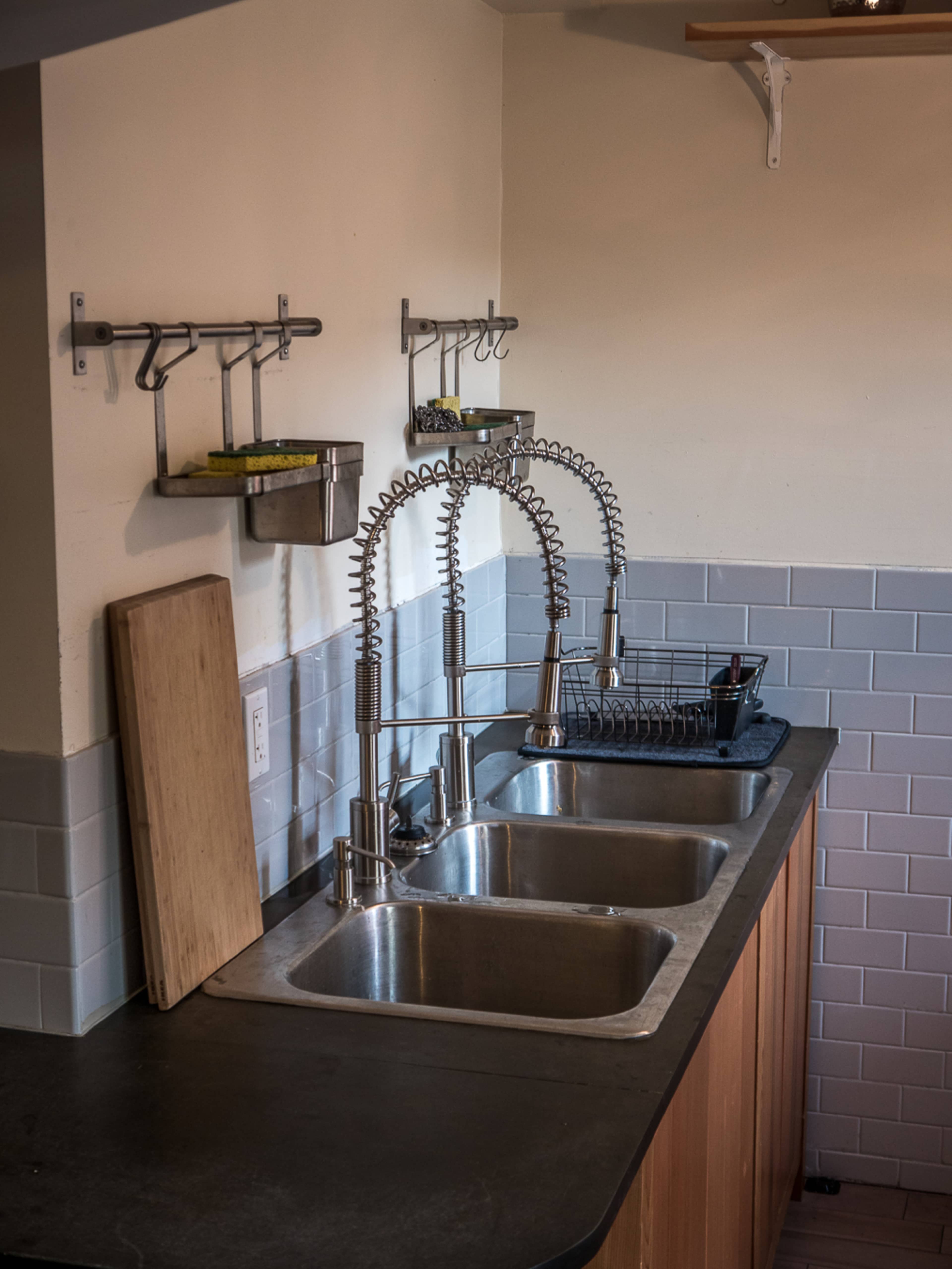 The image shows a kitchen with two stainless steel sinks, a wooden cutting board, and various utensils mounted on the wall.
