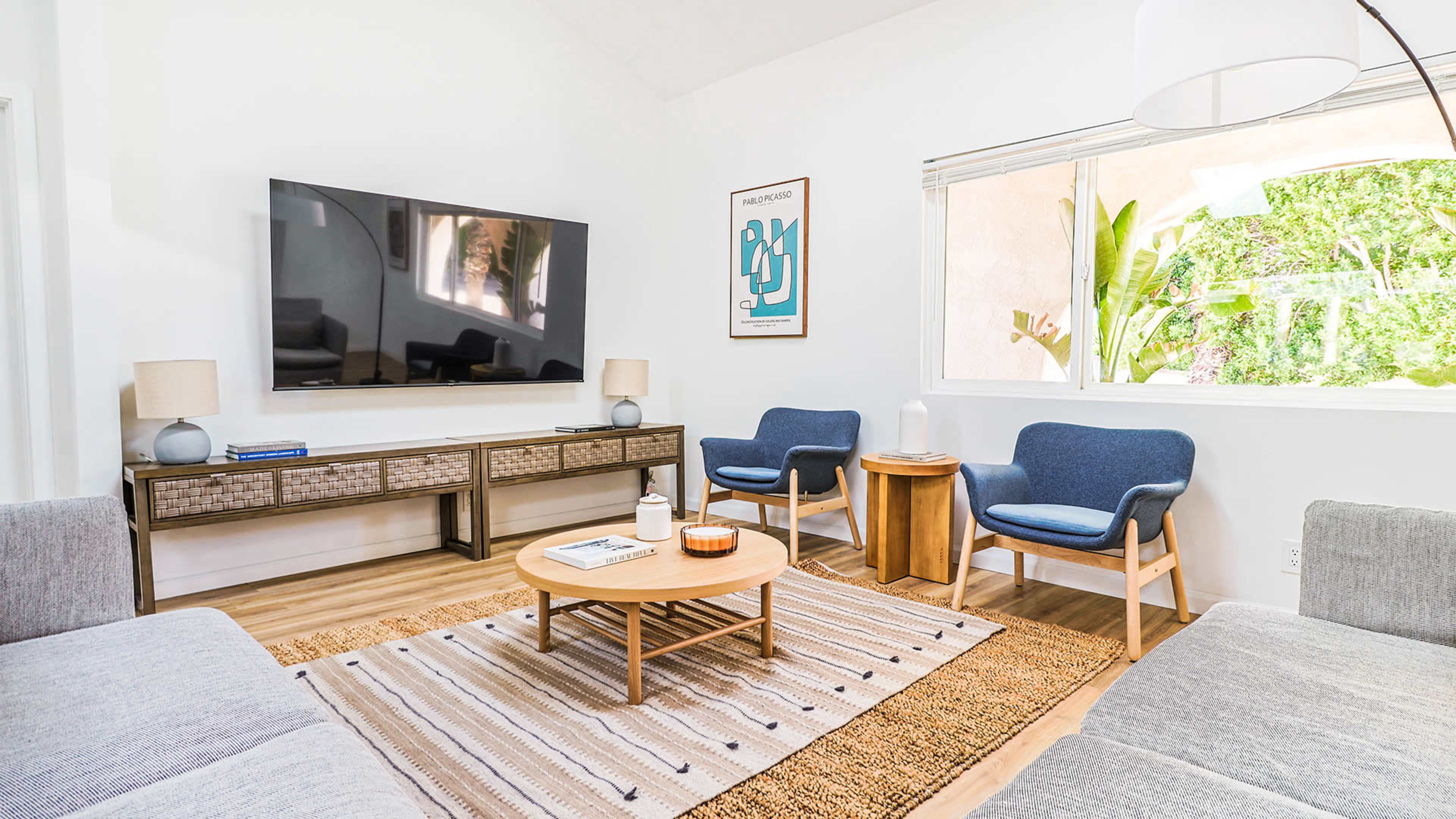 A modern living room featuring a television mounted on the wall, two blue chairs, a circular coffee table, and natural light coming through a window.