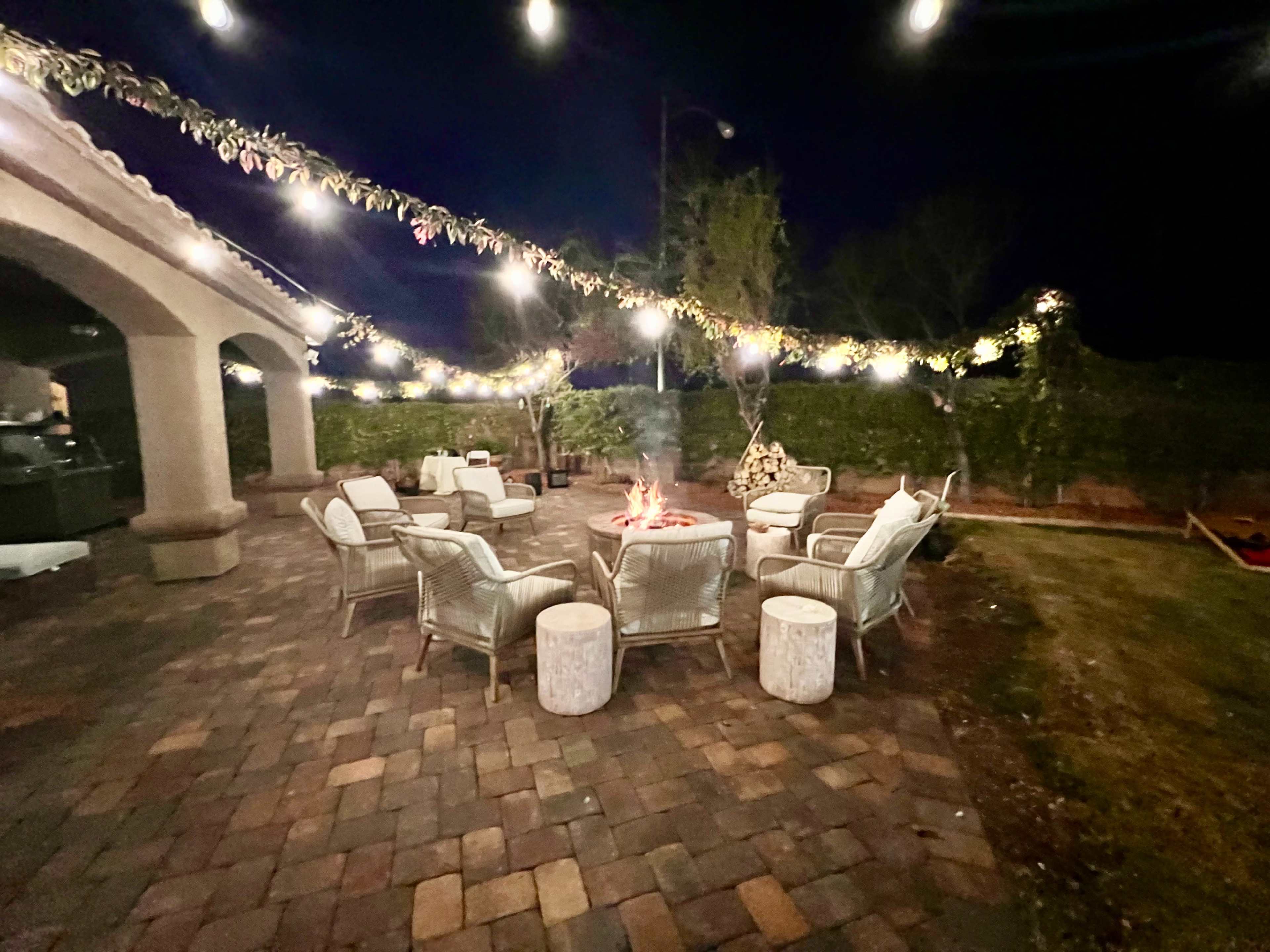 A circular seating arrangement with outdoor chairs surrounds a fire pit, illuminated by string lights in a landscaped backyard at night.