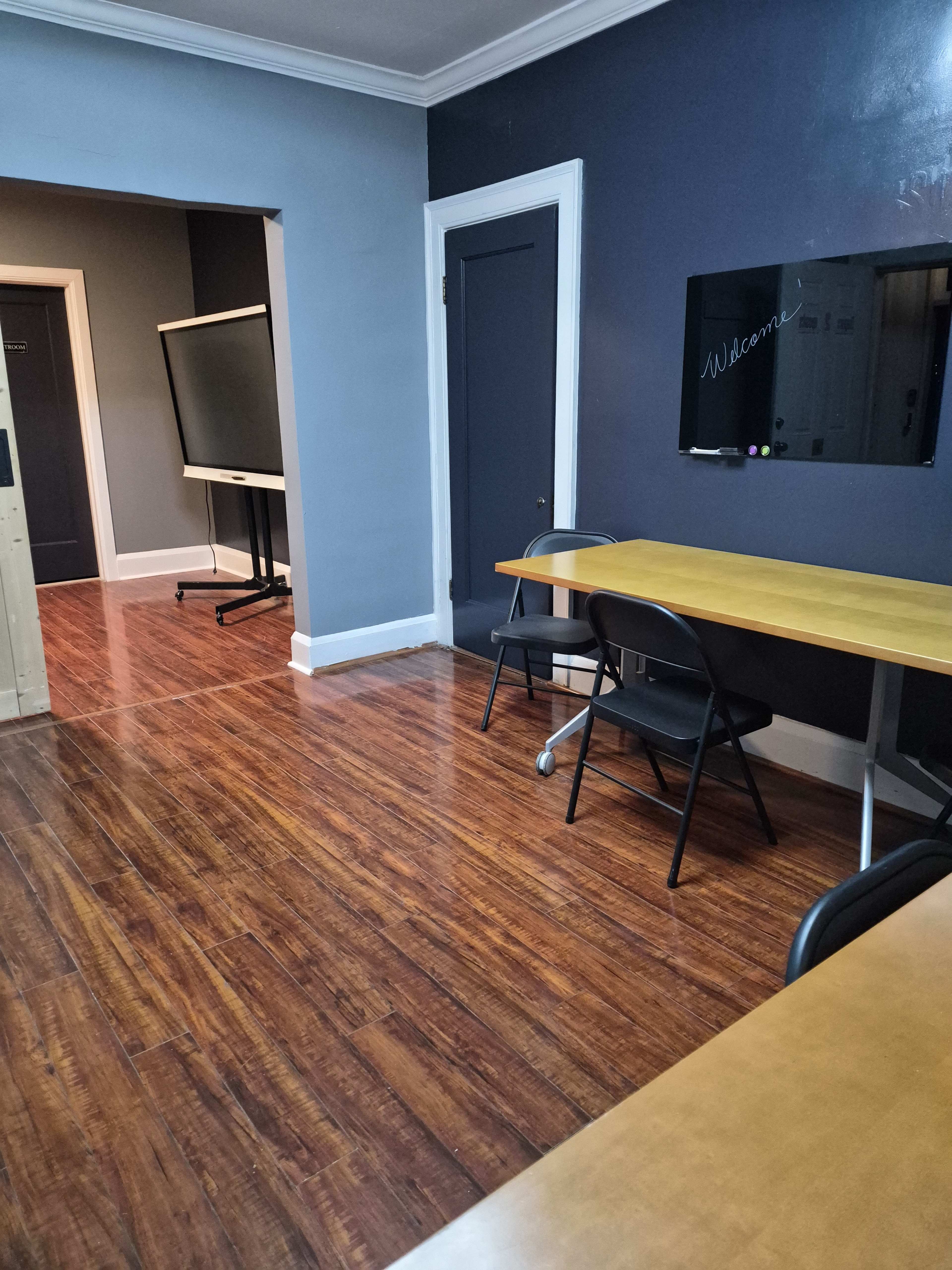 The image shows a modern office space with wooden floors, a table and chairs, and a wall-mounted screen.