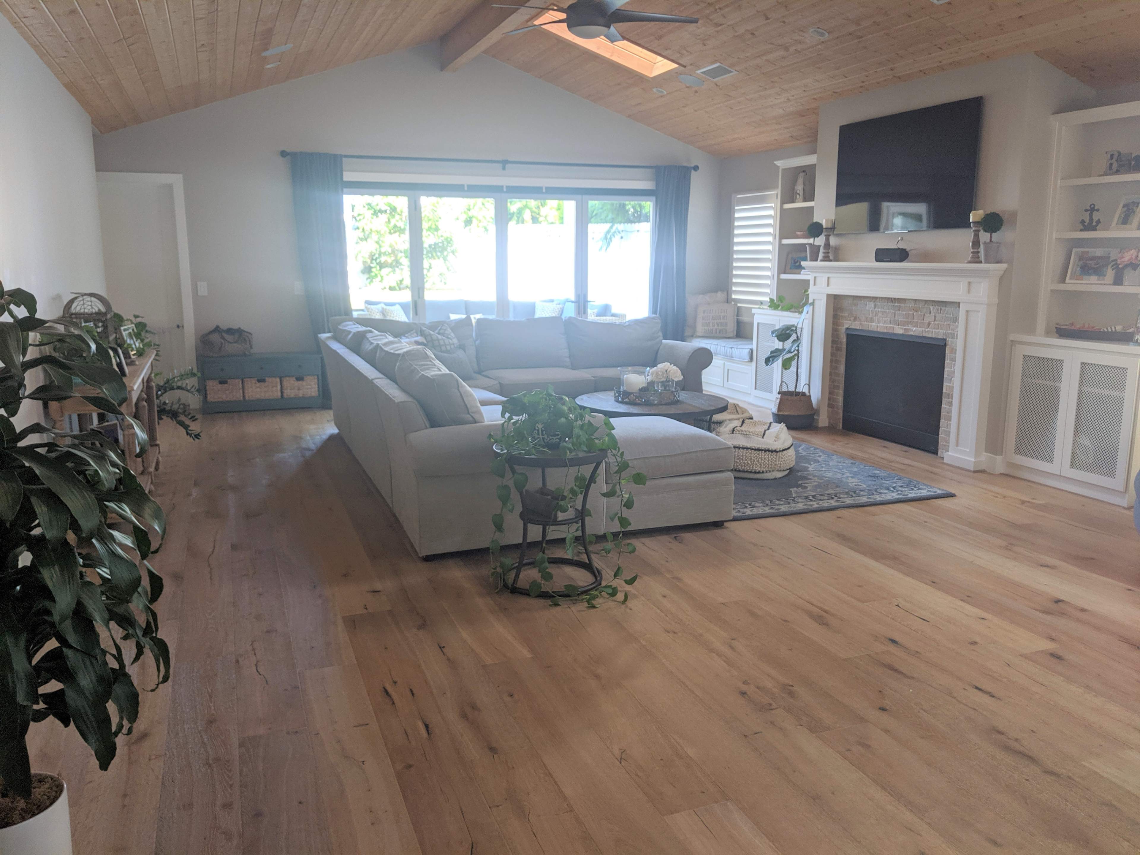 A spacious living room with a light-colored sectional sofa, a coffee table, and large windows letting in natural light, along with a fireplace and wooden ceiling beams.
