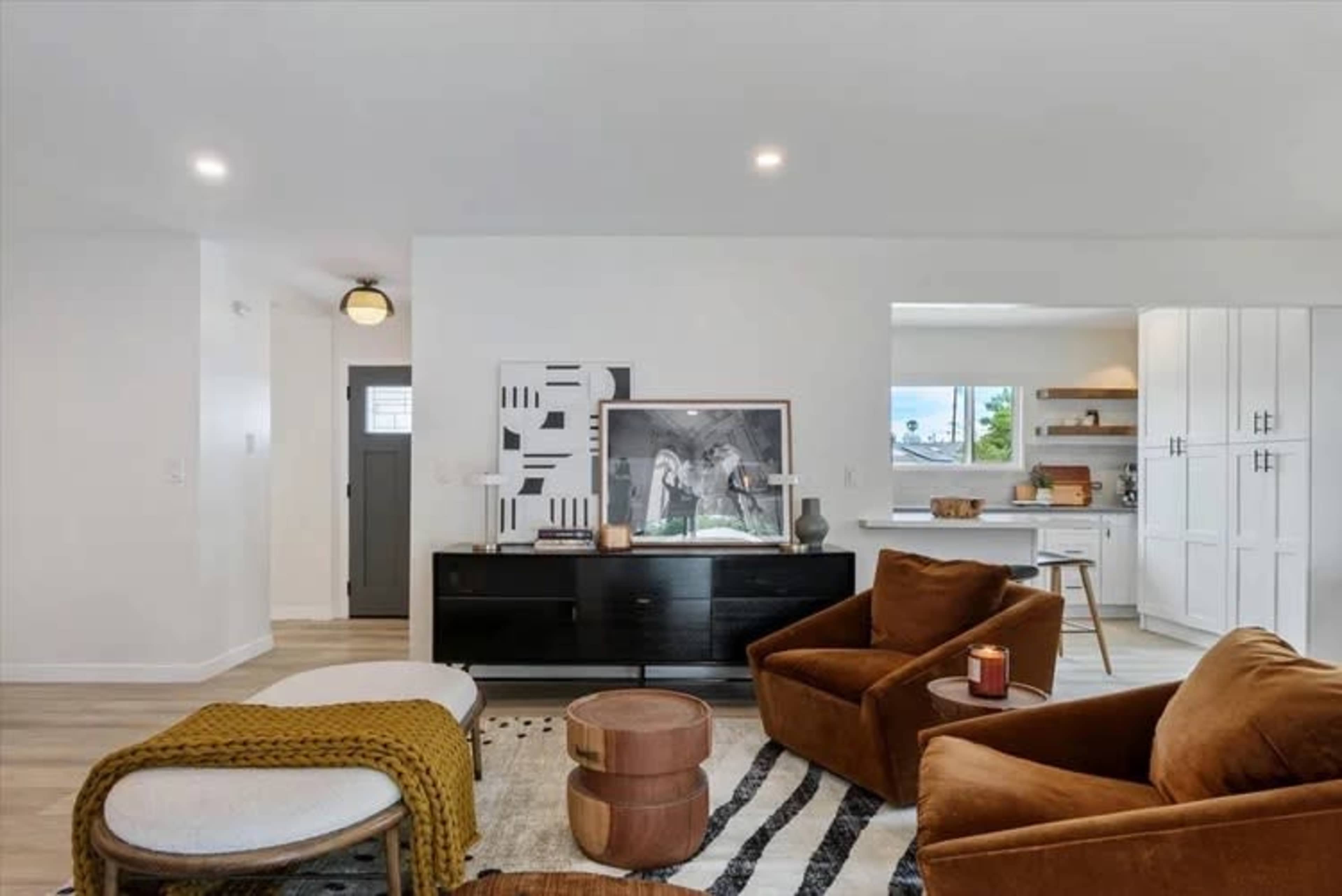 The image shows a modern living room with brown armchairs, a black console table, a large artwork on the wall, and a kitchen area visible in the background.