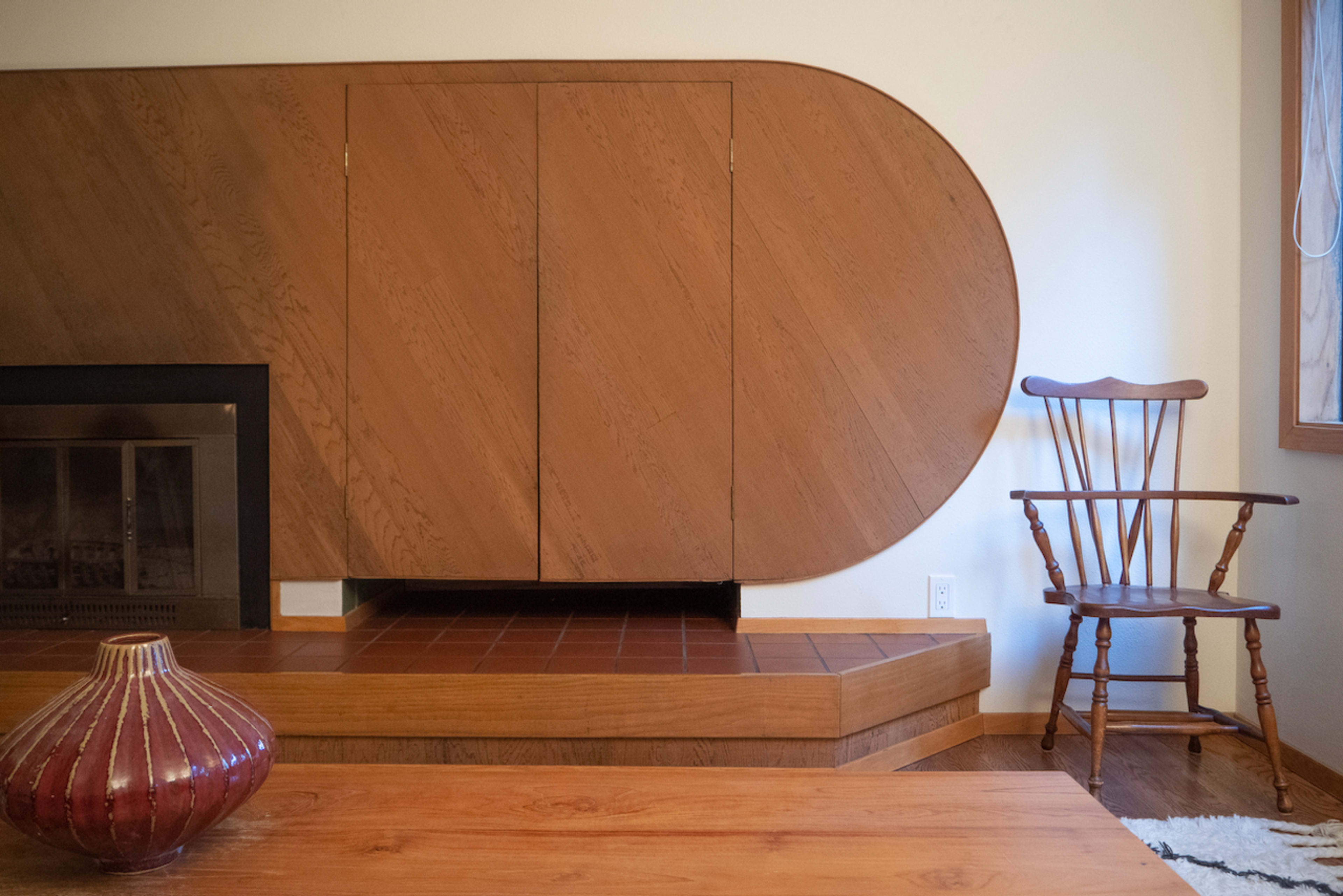 A modern interior featuring a curved wooden wall cabinet, a fireplace, a wooden chair, and a low wooden table.