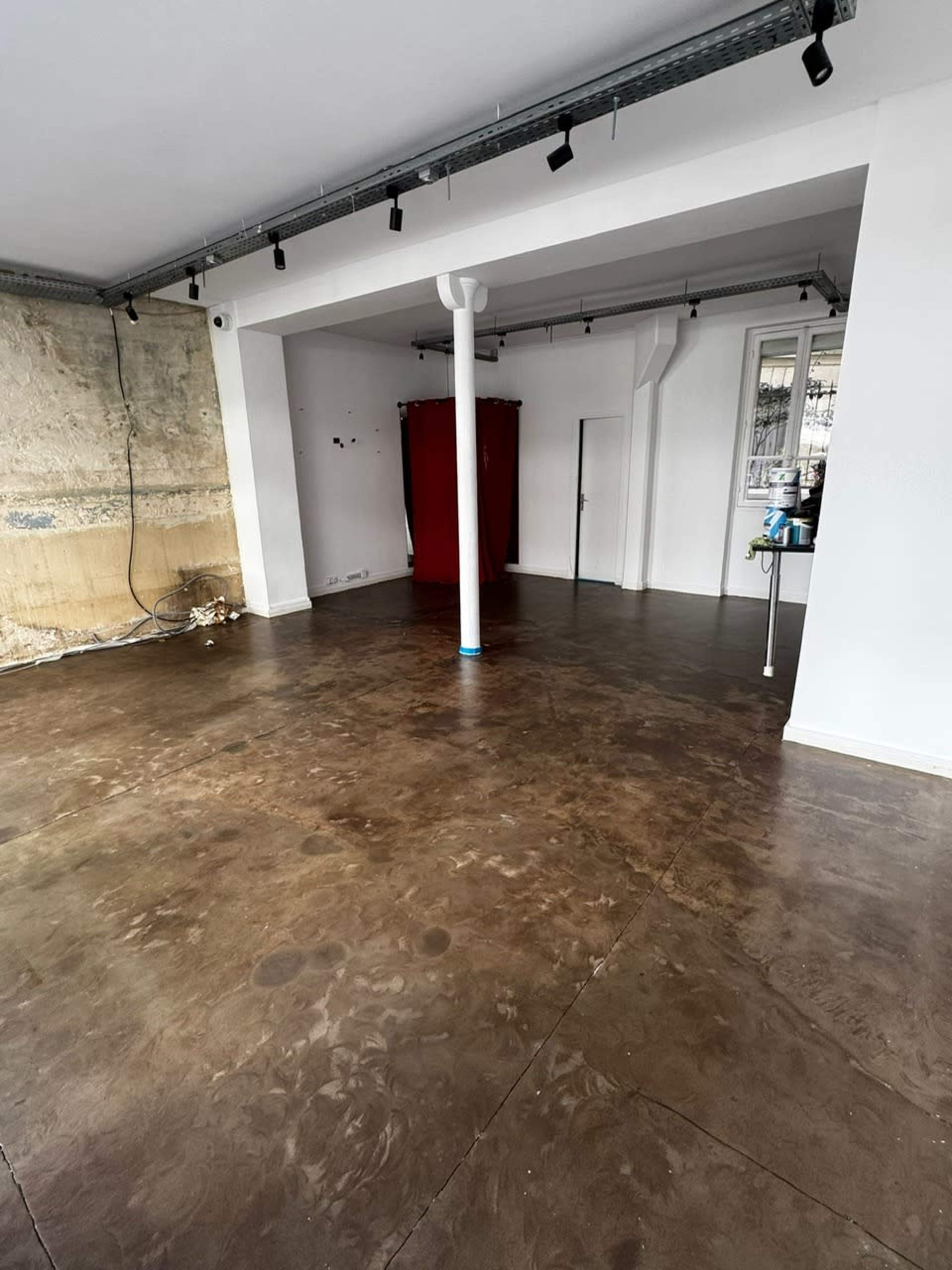 The image shows an empty room with polished concrete flooring, white walls, a central support column, and a red curtain in the background.