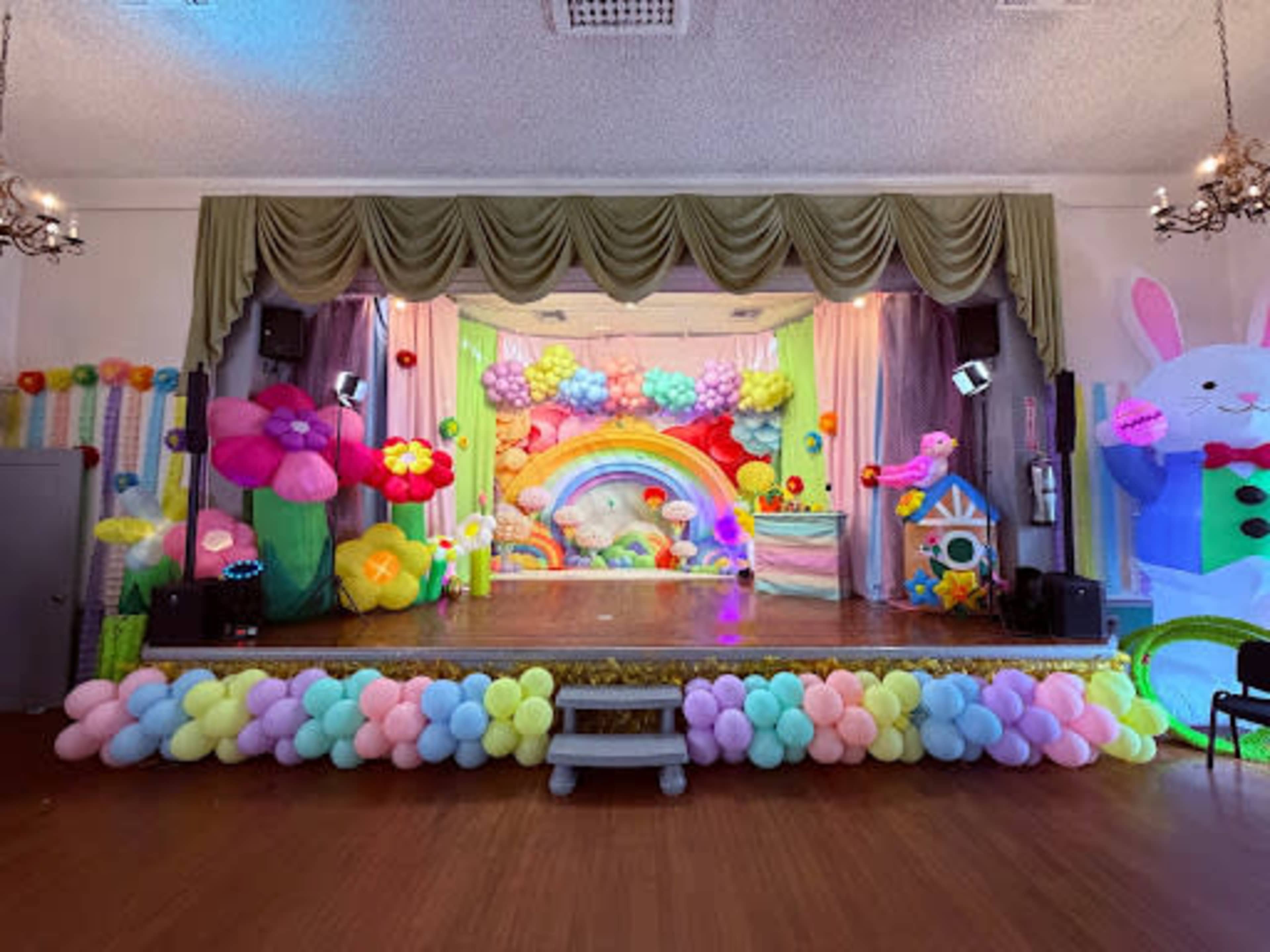 The scene depicts a colorful stage decorated with balloons, flowers, and a rainbow backdrop, intended for a festive event.