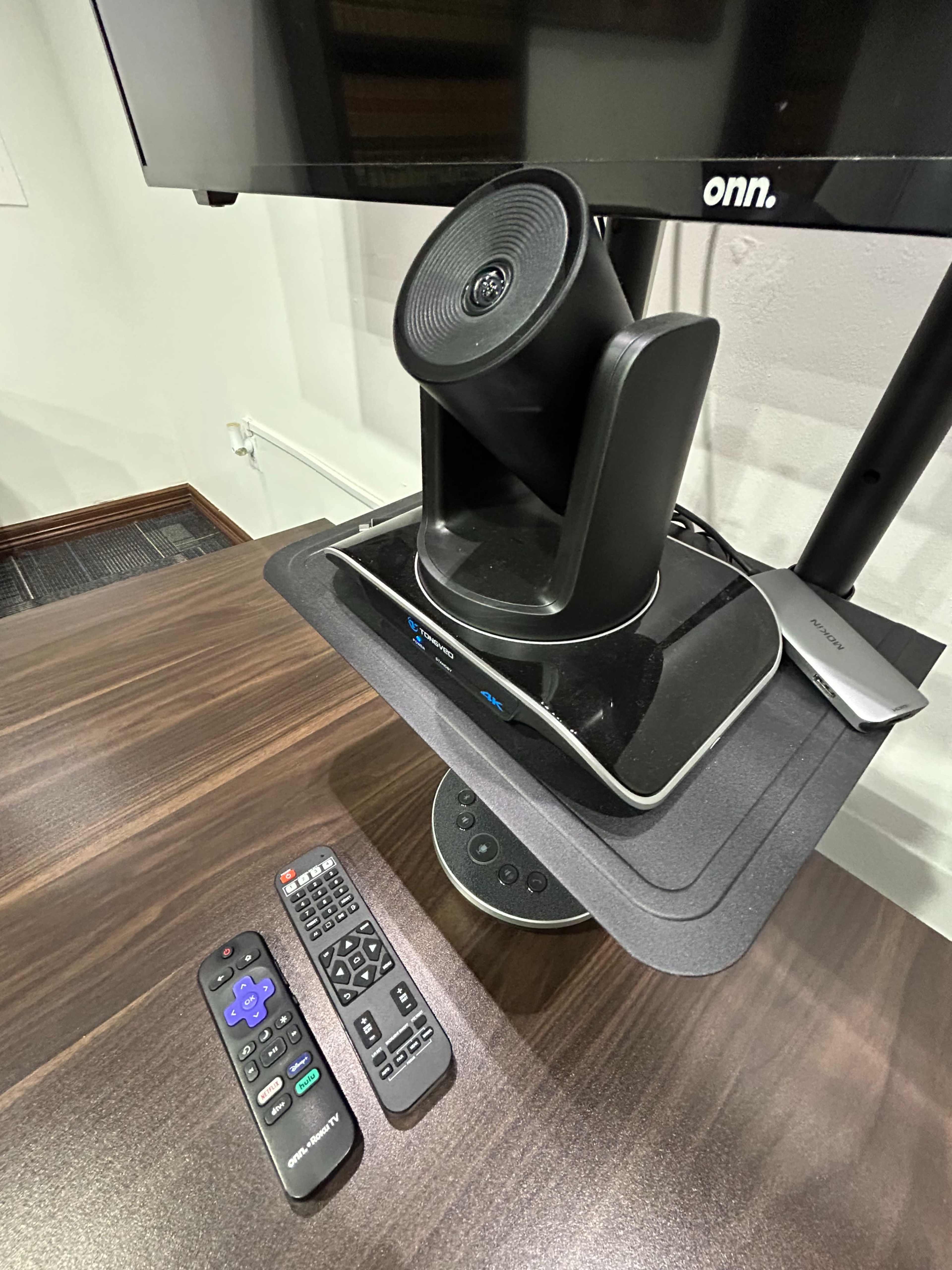 A black speaker is mounted on a stand beneath a television, next to two remote controls on a dark wooden surface.