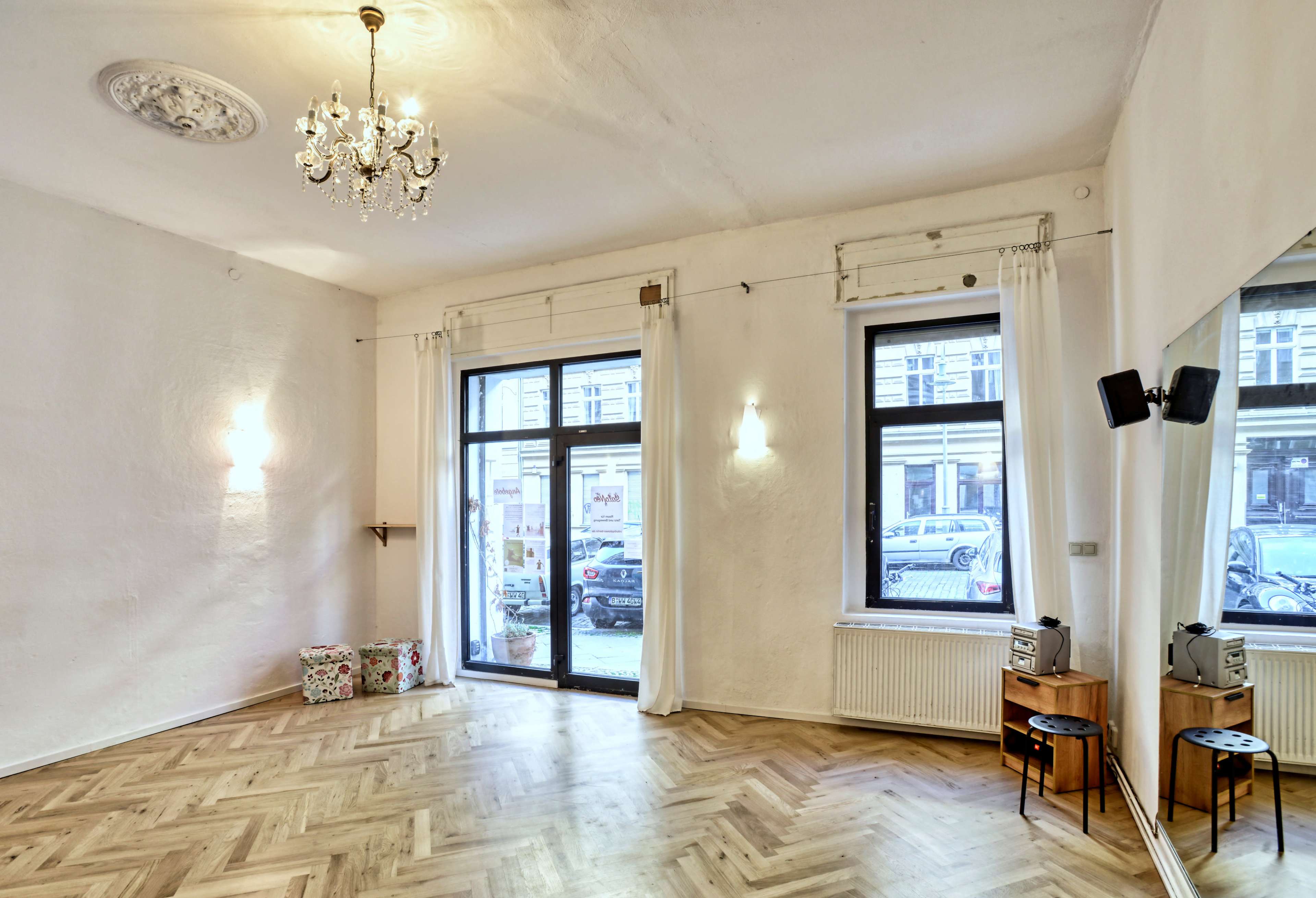 The room features wooden flooring, large windows with sheer curtains, and a chandelier overhead, with two small tables and colorful boxes in one corner.