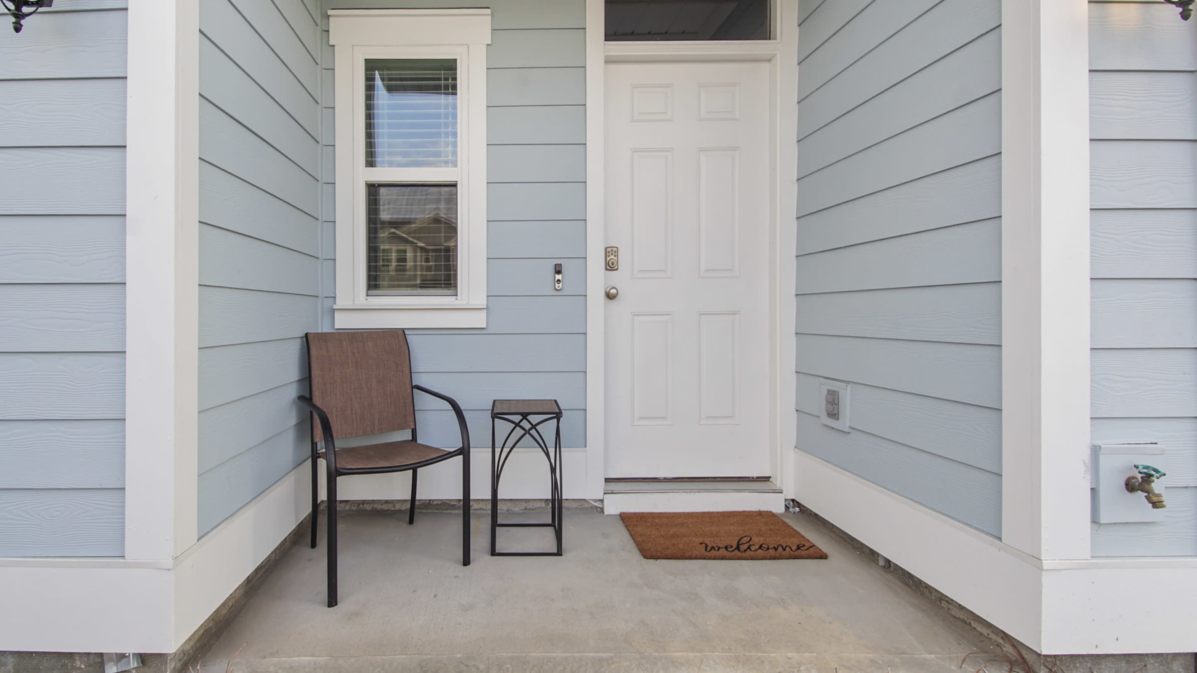 The image shows a front porch with a chair, a small table, and a welcome mat in front of a white door on a light blue house.