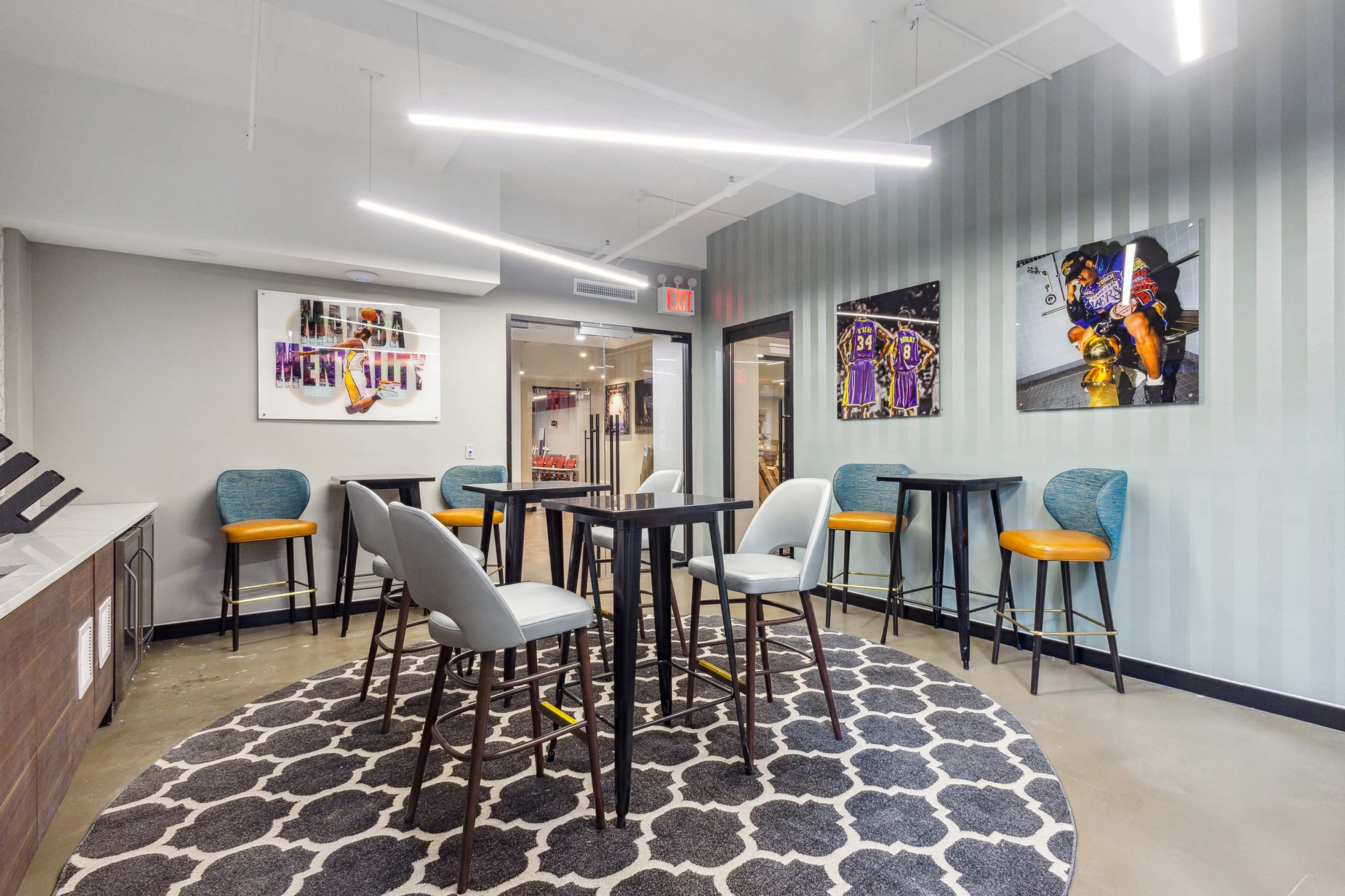 The image shows a modern lounge area with several high tables and chairs arranged on a patterned rug, alongside colorful wall art.
