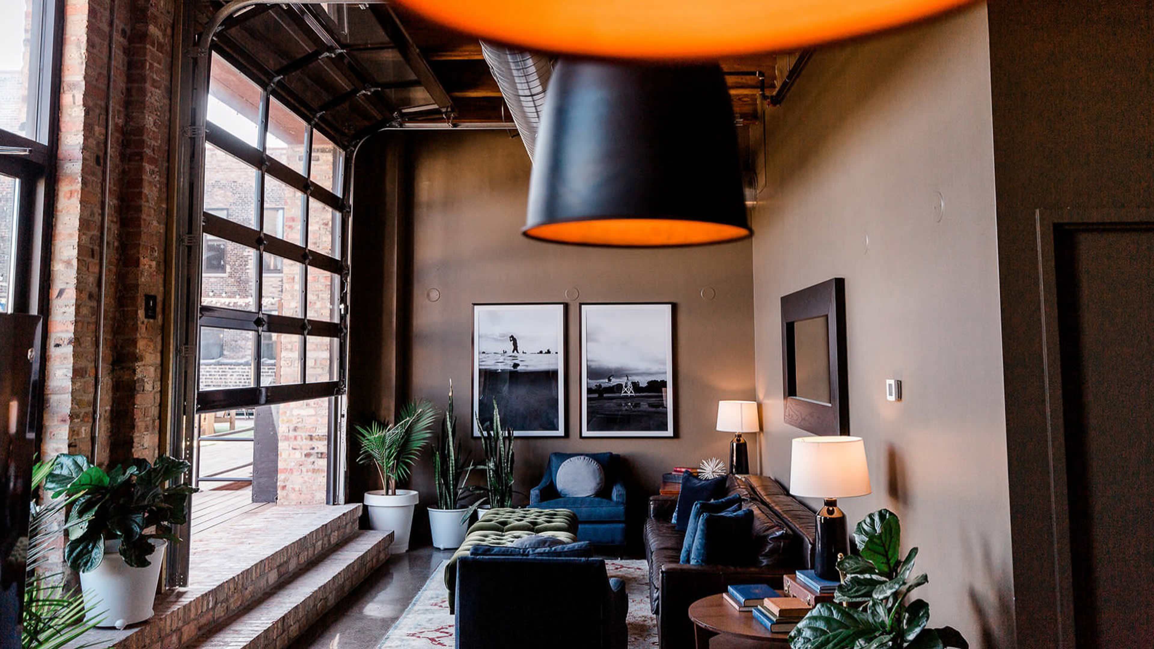 The image shows a modern living room with large windows, two framed photographs on the wall, a green upholstered ottoman, and several indoor plants.