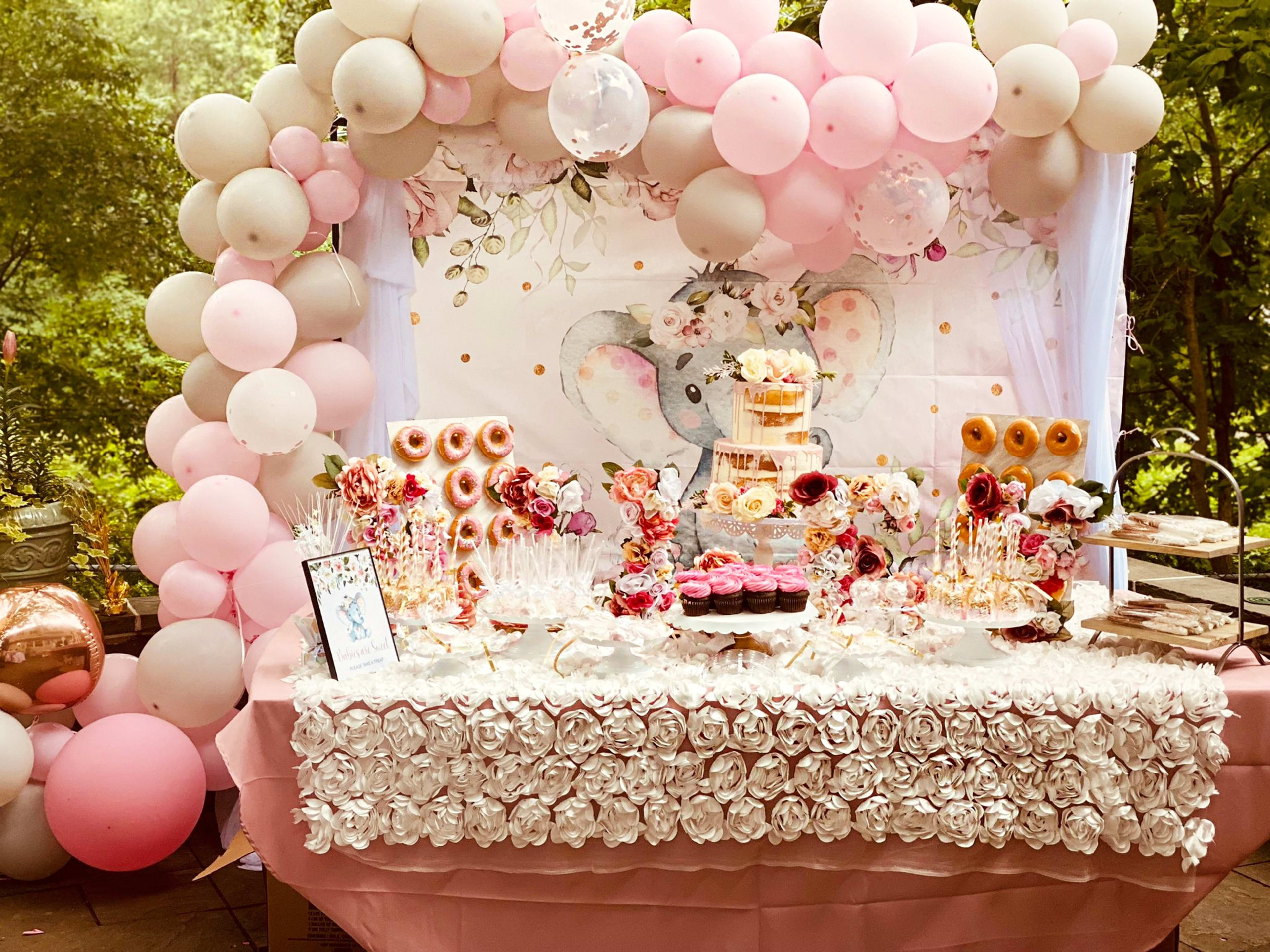 A pink and white cake sits atop a table decorated with balloons for a girl's outdoor baby shower.