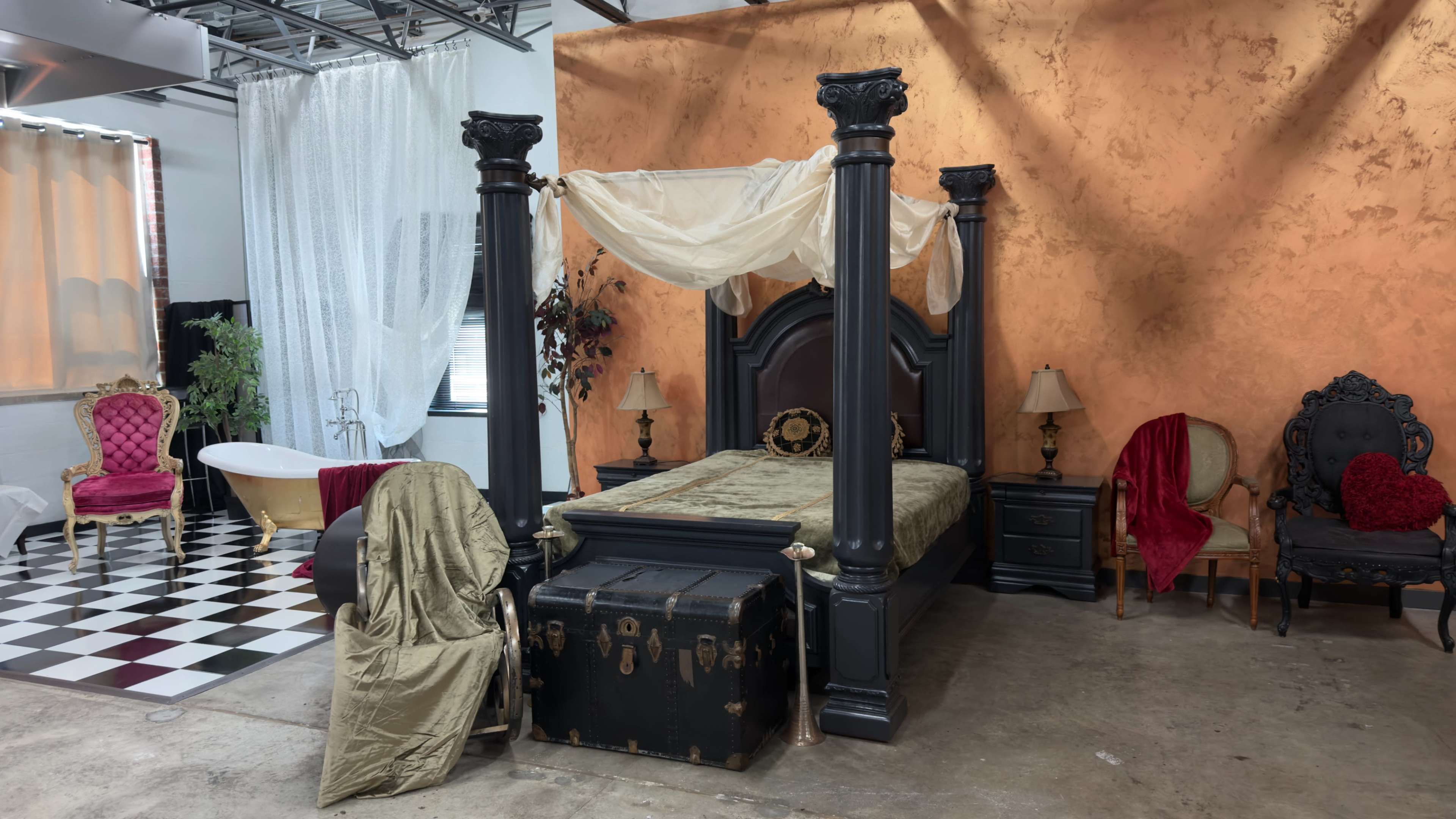 The image shows a styled interior featuring a four-poster bed with a draped canopy, a vintage trunk at its foot, and various seating arrangements nearby, all set against an orange wall.