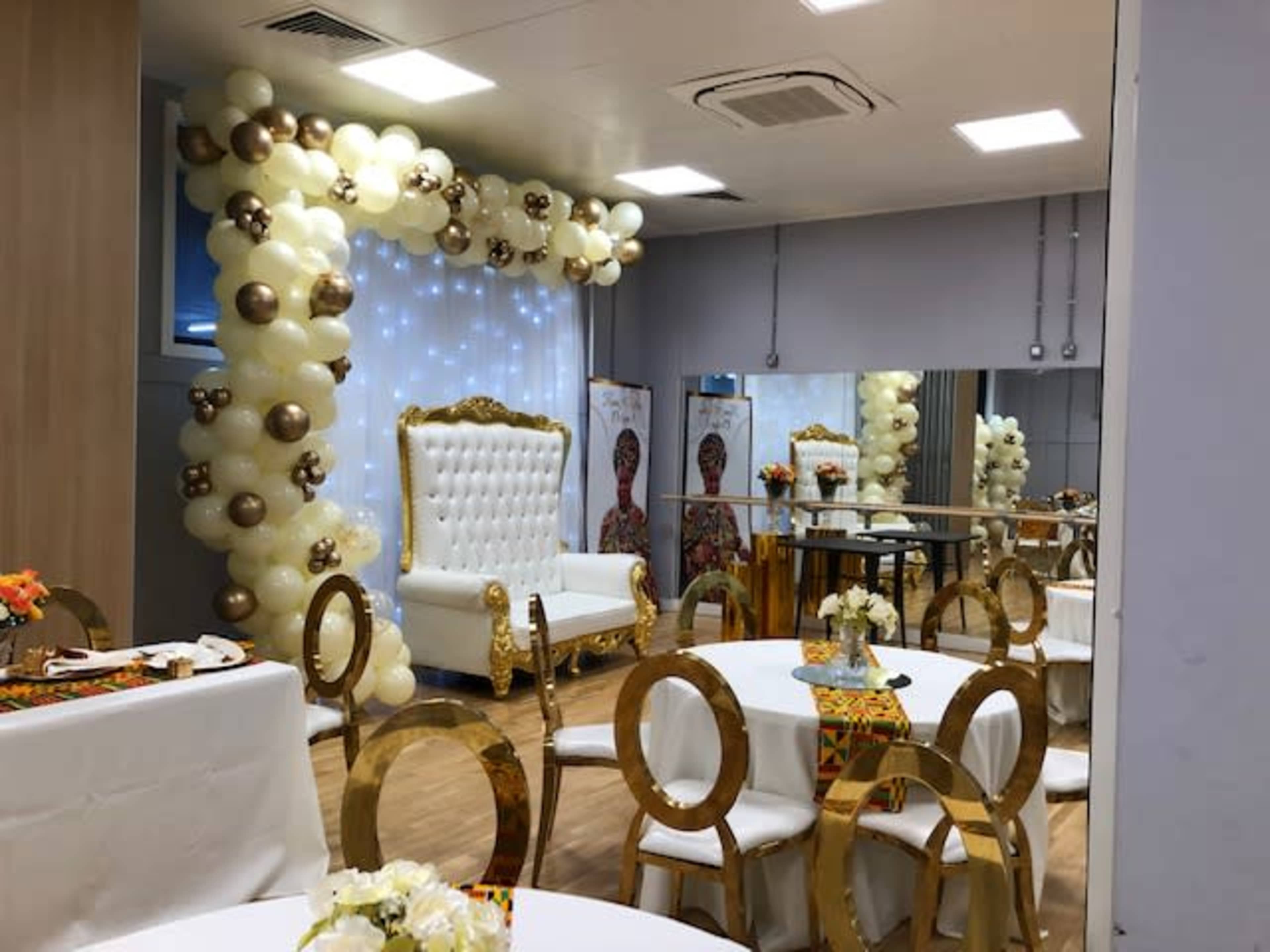 A decorated event space featuring a white upholstered throne chair, round tables with flower centerpieces, and a backdrop of balloons in gold and cream.