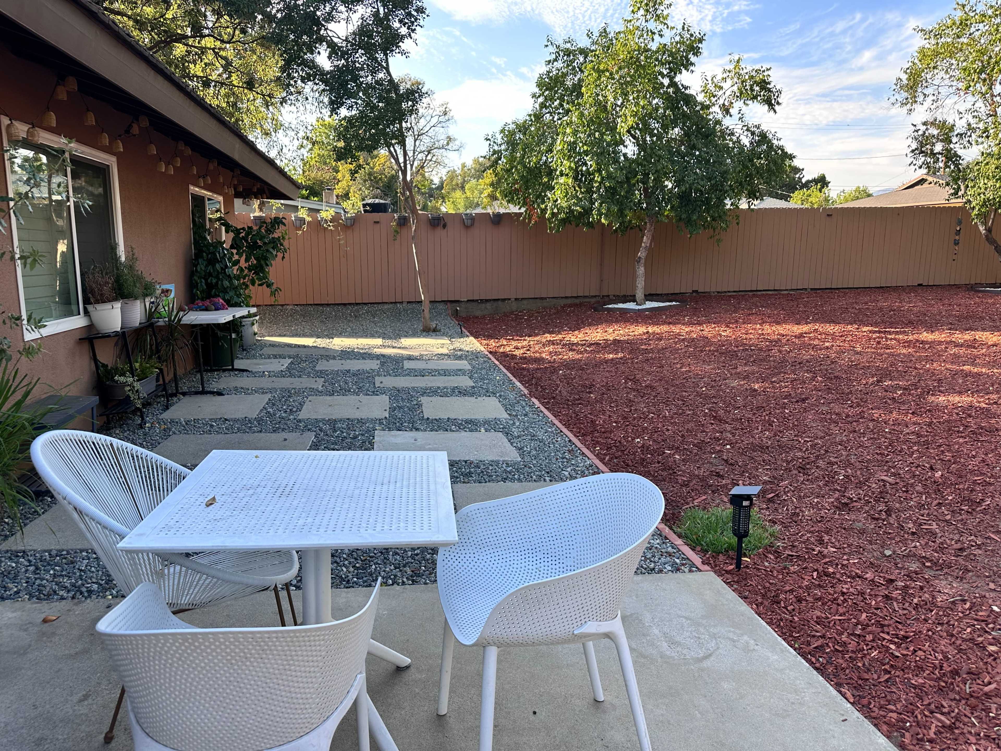 A small outdoor patio features a white table and chairs, surrounded by gravel pathways and red mulch, with trees and a fenced area in the background.