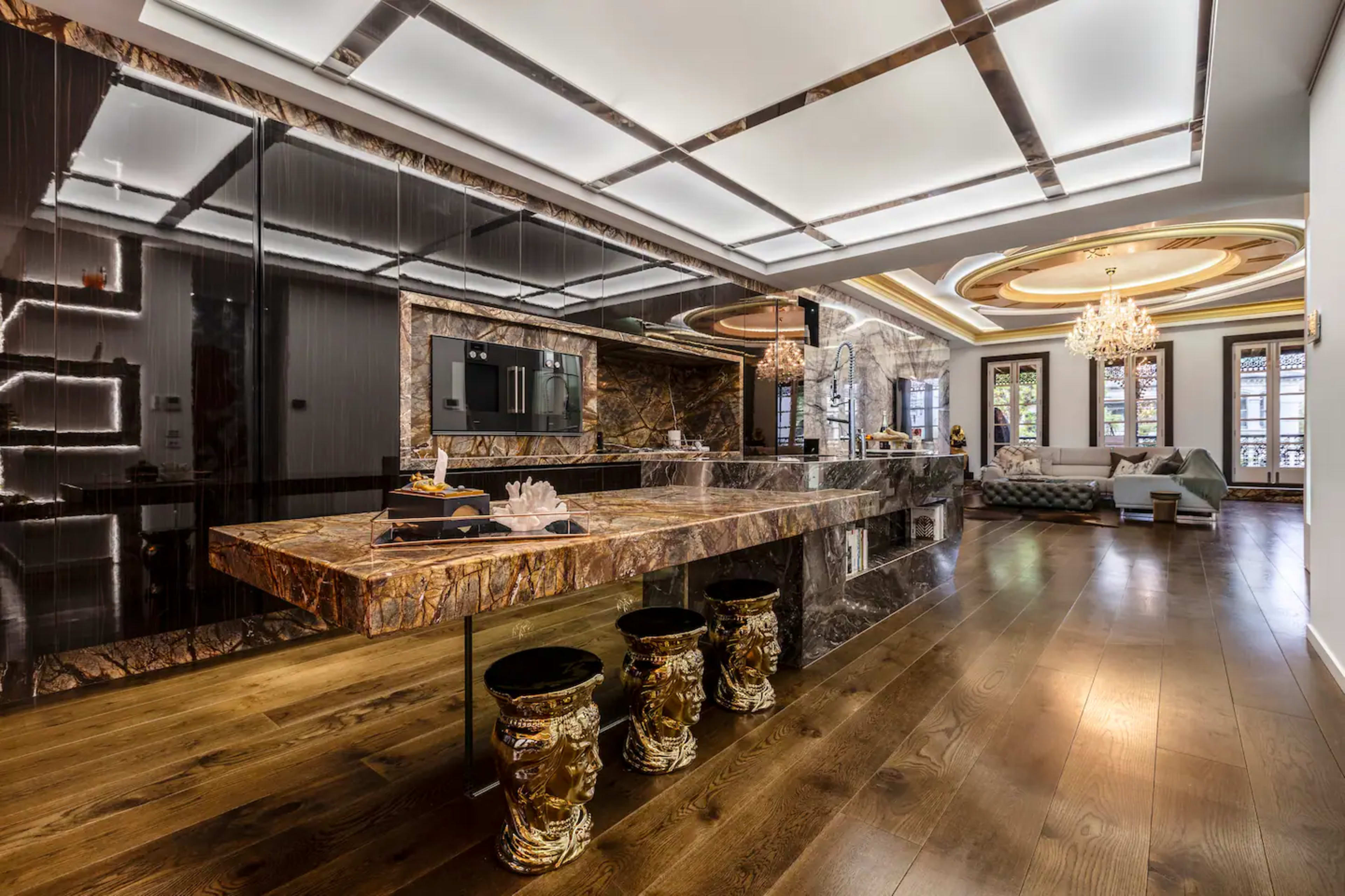 A modern kitchen with a large marble island, sleek cabinets, chandeliers, and a spacious living area in the background.