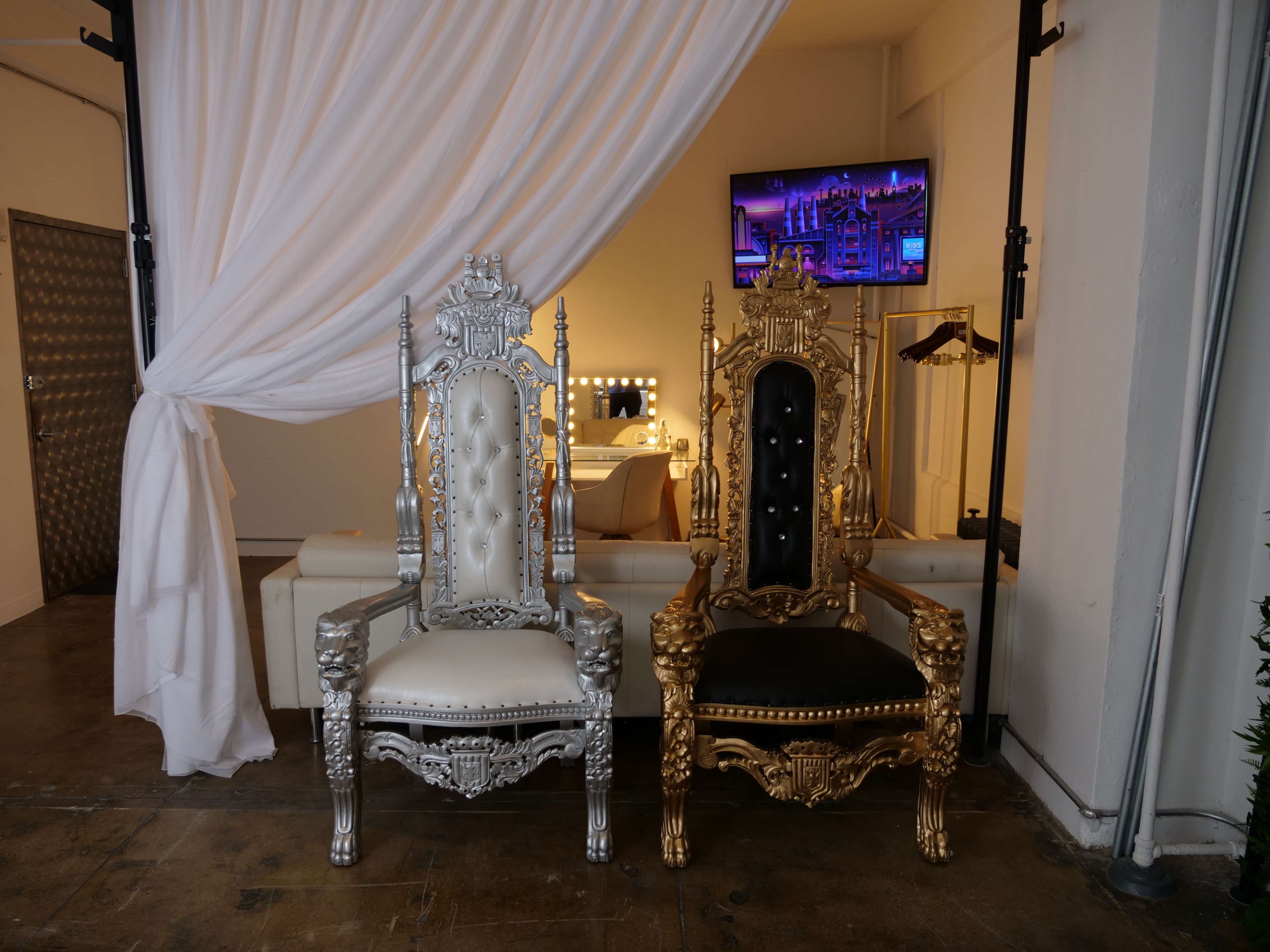Two ornate chairs, one silver and white and the other gold and black, placed against a backdrop of a white draped curtain in a spacious room.