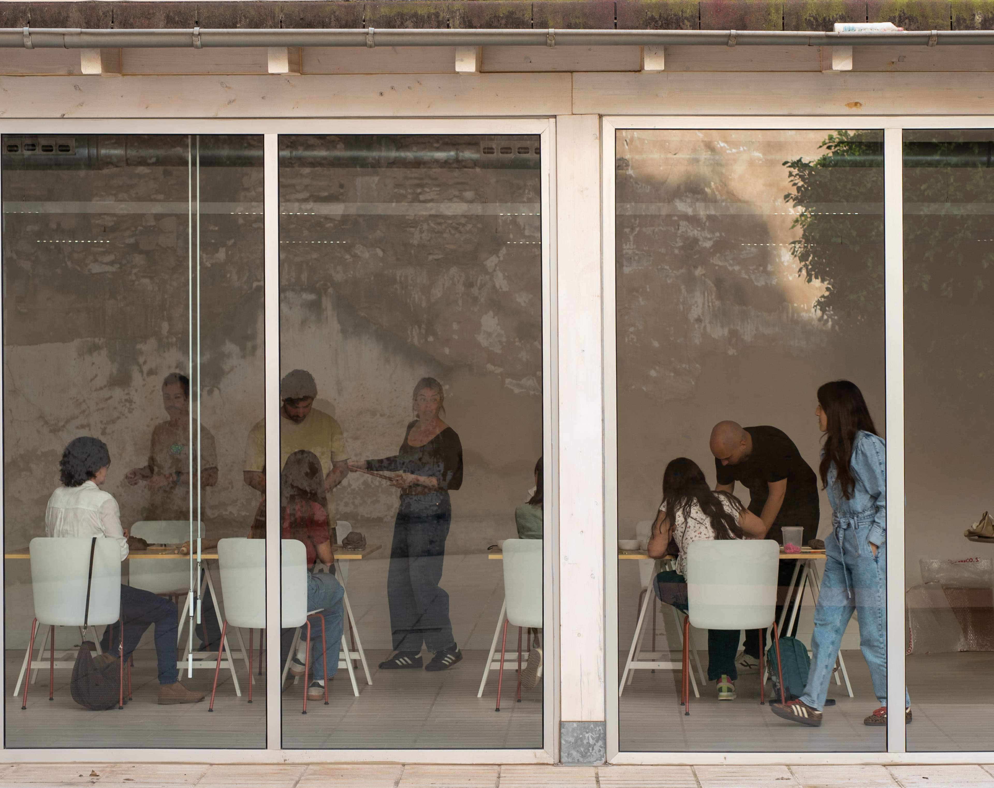 People are gathered around tables inside a modern room with large glass windows, engaging in an activity or discussion.