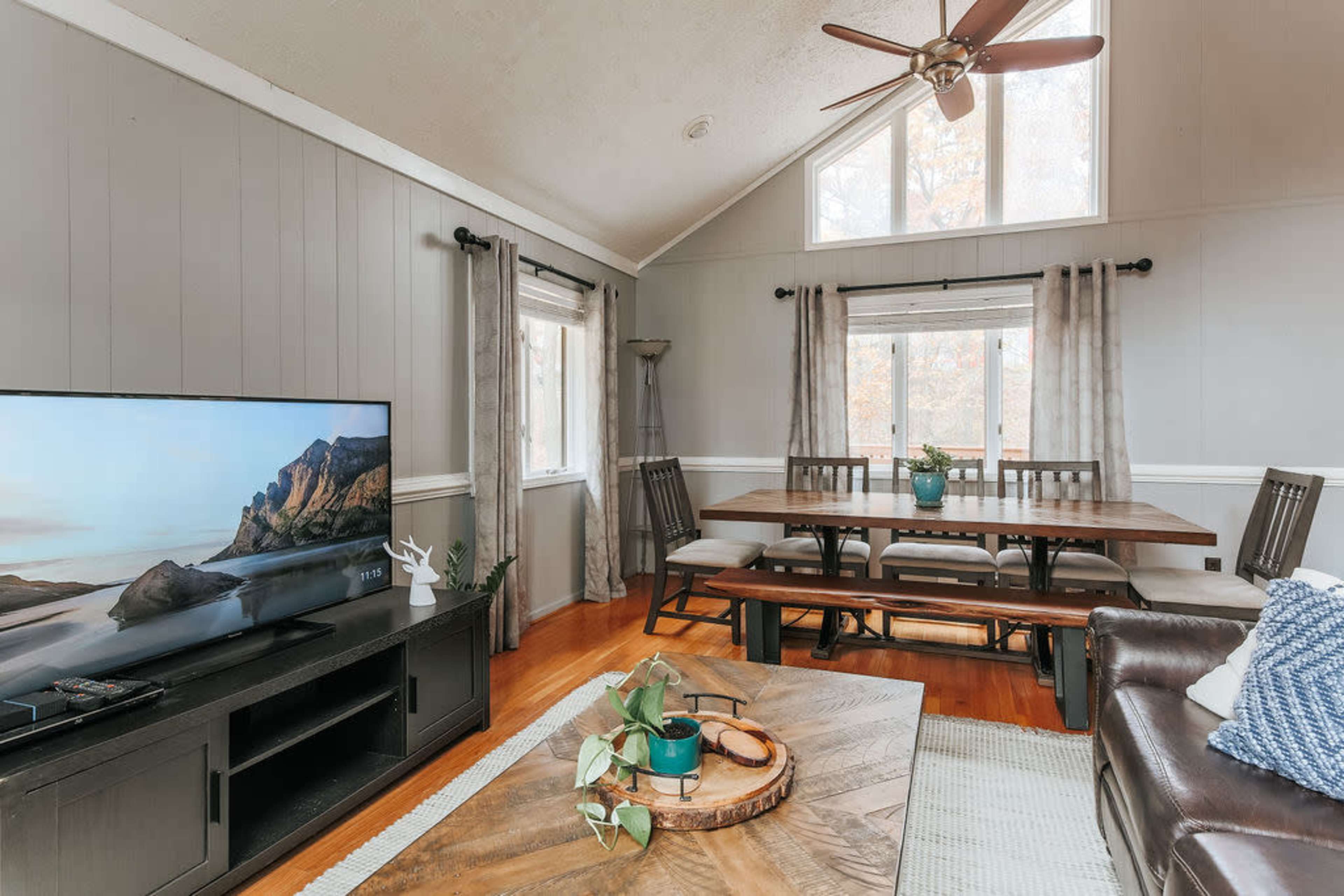 A living room features a large television, a dining table, and two windows providing natural light, with a ceiling fan overhead.
