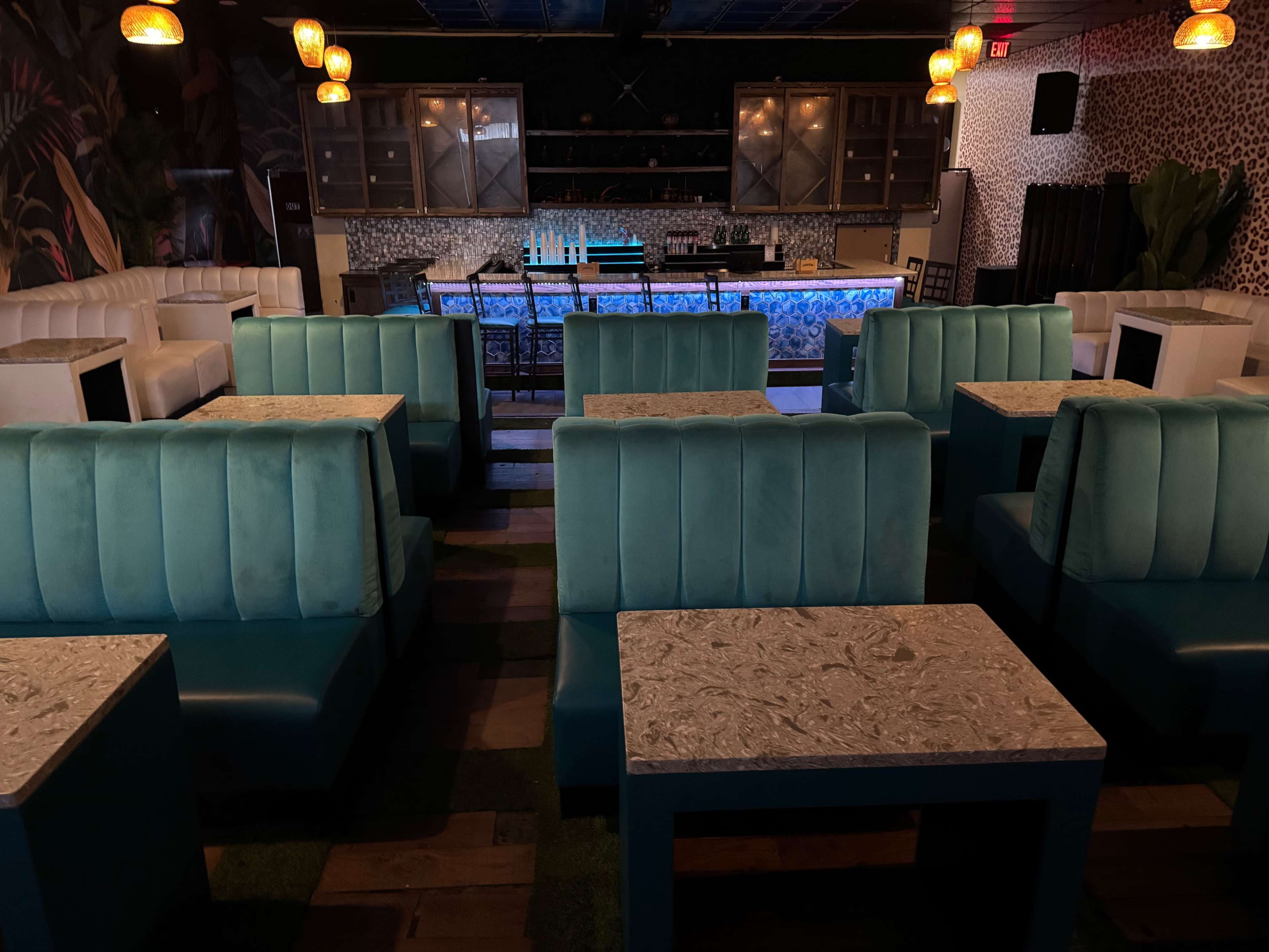 The image shows an empty restaurant interior with teal booths and marble-top tables arranged around a bar area illuminated by pendant lights.