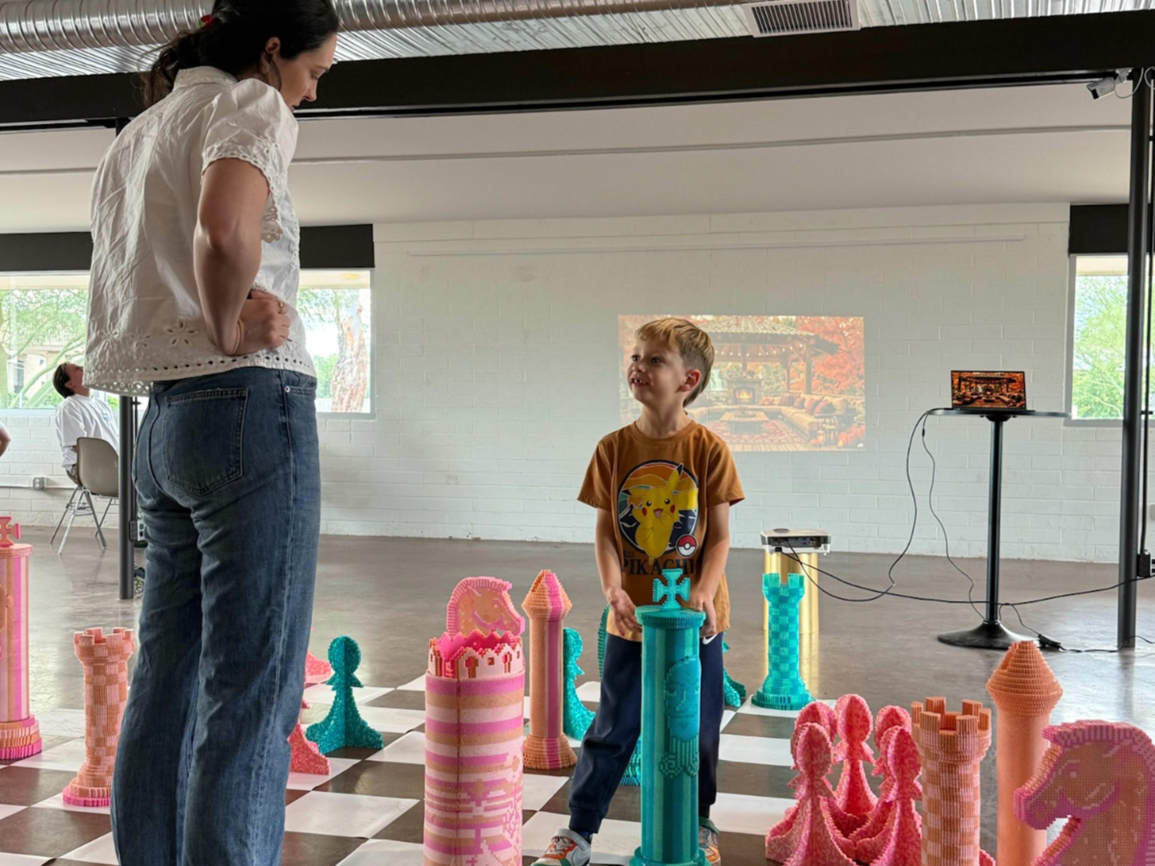 A woman and a young boy stand in an art installation featuring oversized, colorful chess pieces in a spacious room.