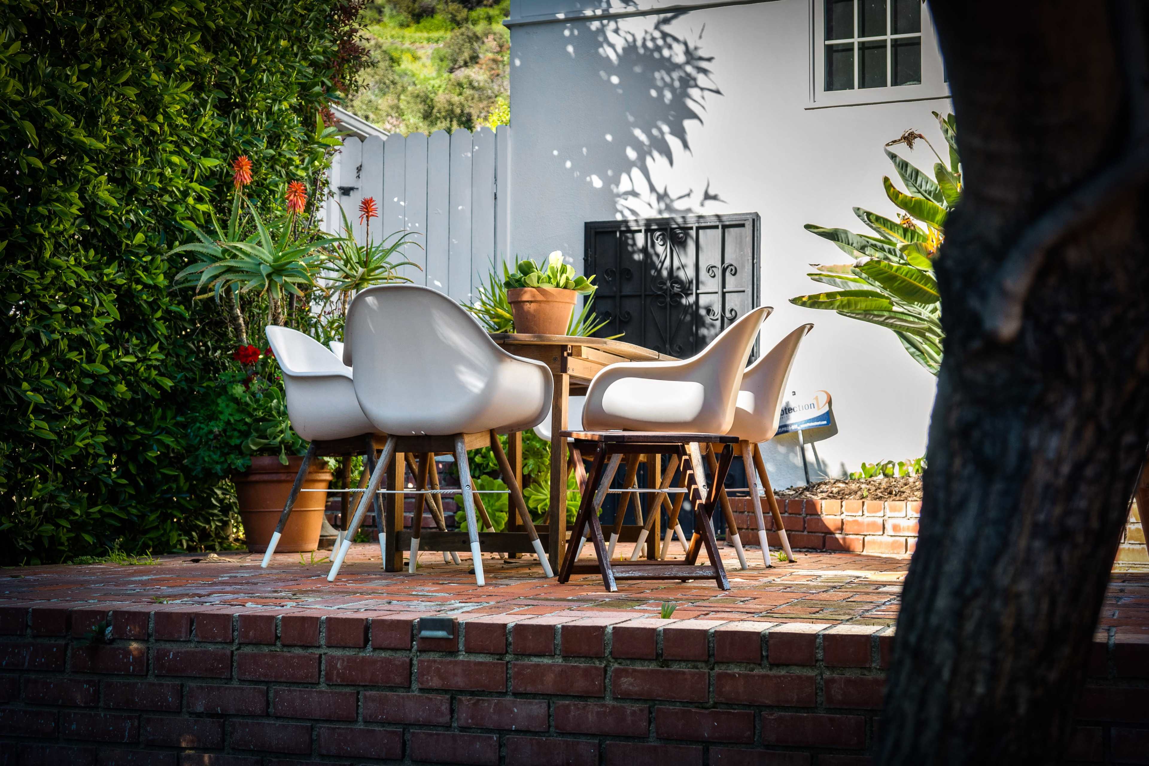 A set of modern chairs surrounds a wooden dining table on a brick patio, with potted plants and greenery in the background.