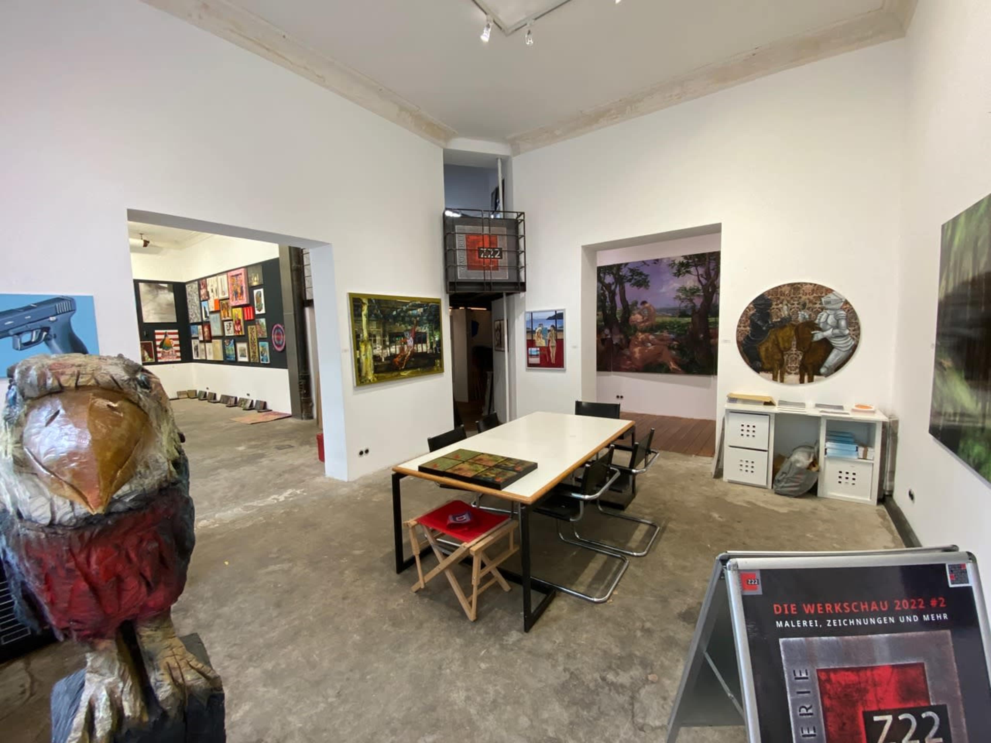 An art gallery interior with a central table surrounded by chairs, featuring various artworks displayed on the walls.
