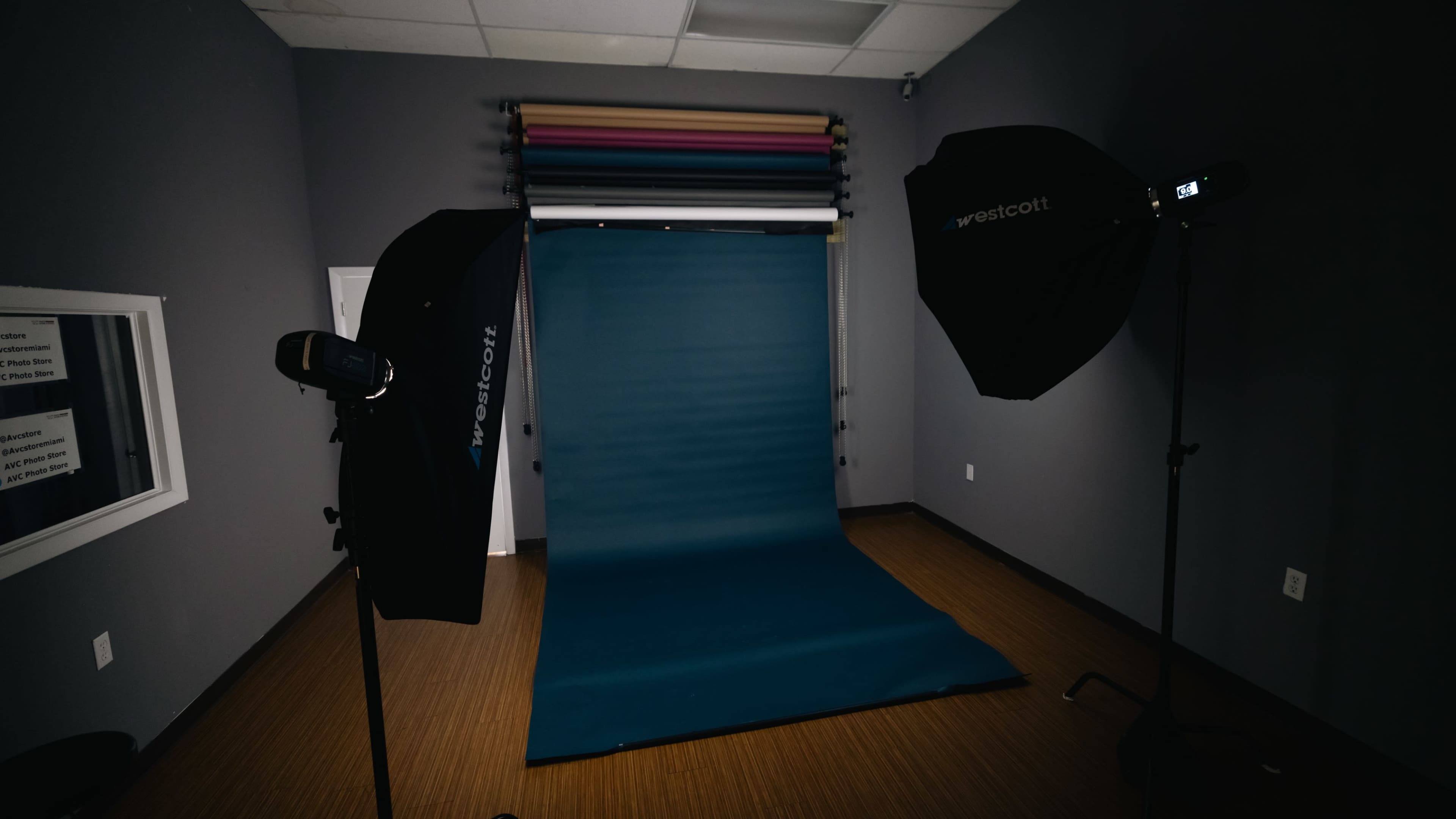 A photography studio setup with a teal backdrop, two softbox lights, and colored paper rolls on a rack.