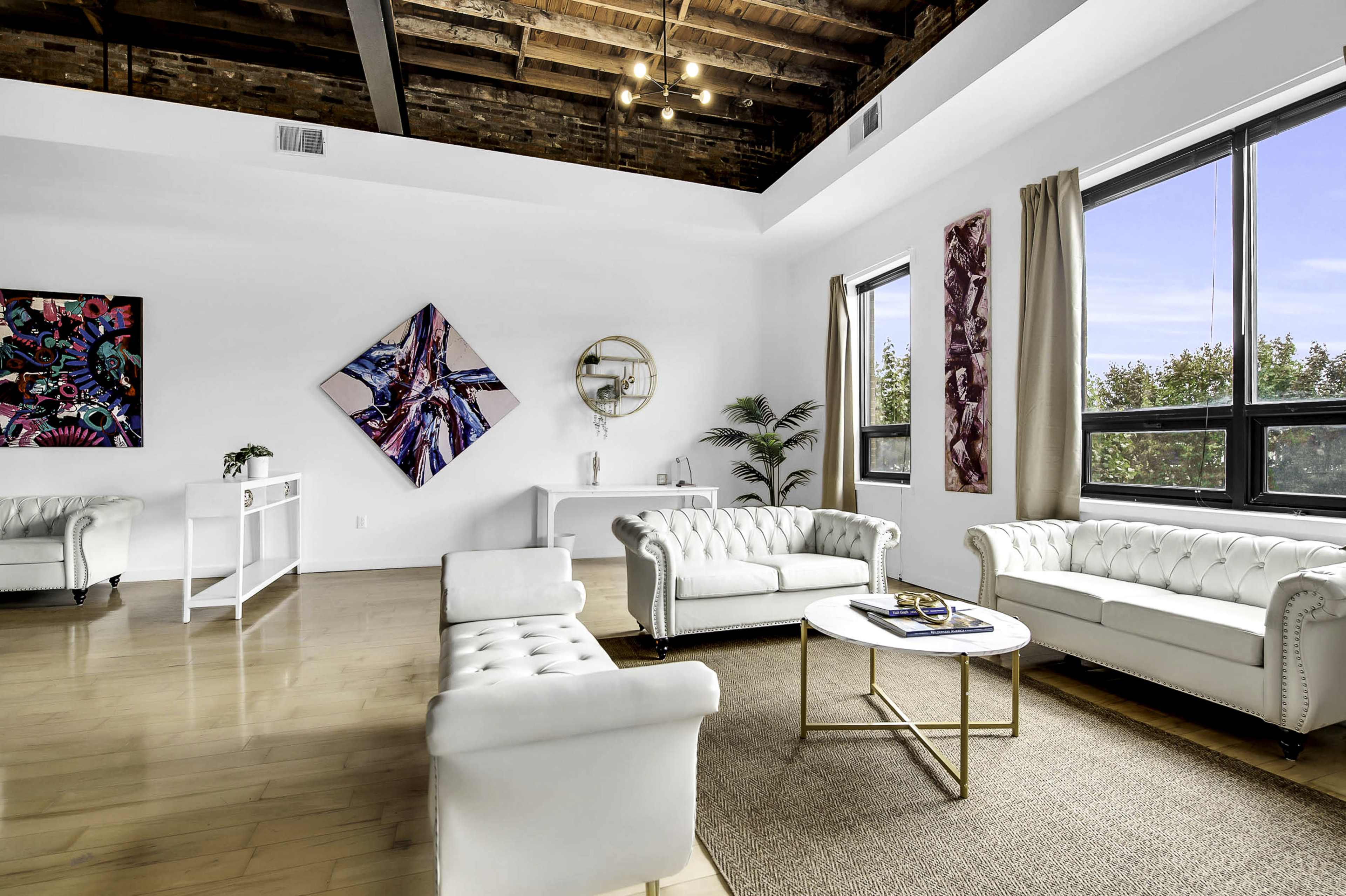A modern living room with white leather sofas, a round coffee table, and large windows allowing natural light in, complemented by contemporary artwork on the walls.