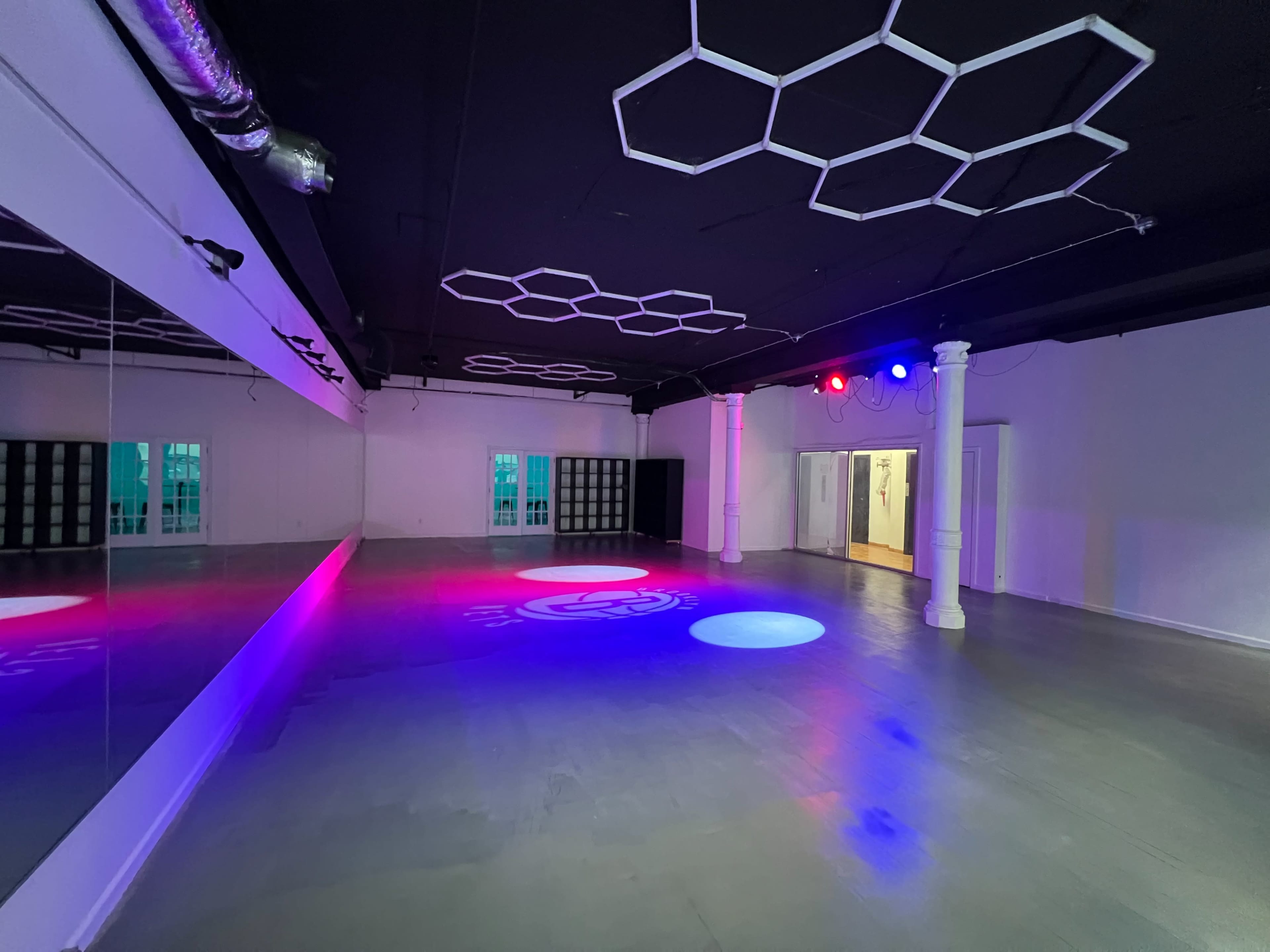 The image shows a spacious, empty dance studio with a reflective mirror wall and colorful lighting fixtures on the ceiling.