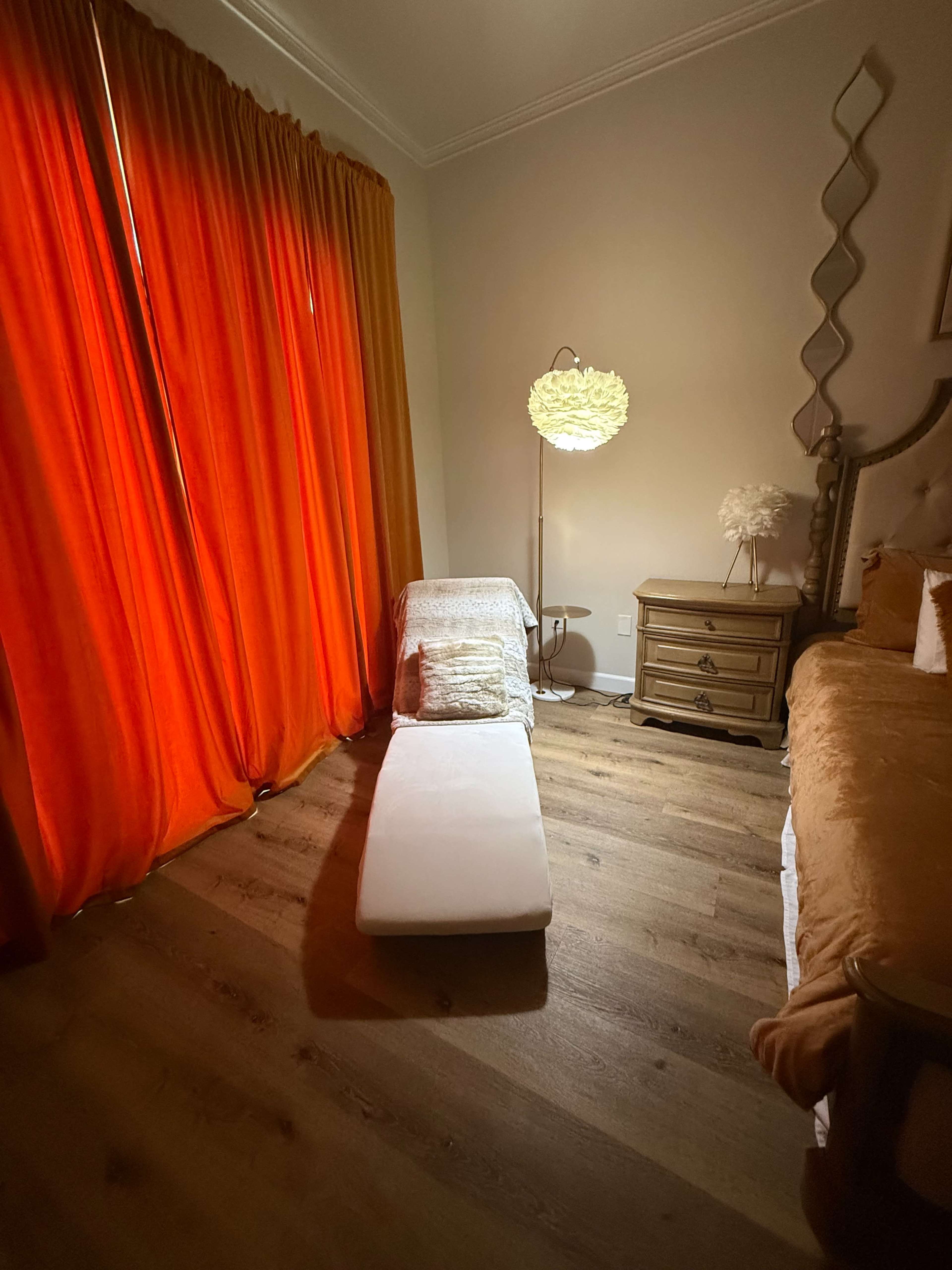 The image shows a cozy bedroom corner with a chaise lounge, a bedside table with a lamp, and orange curtains covering a window.
