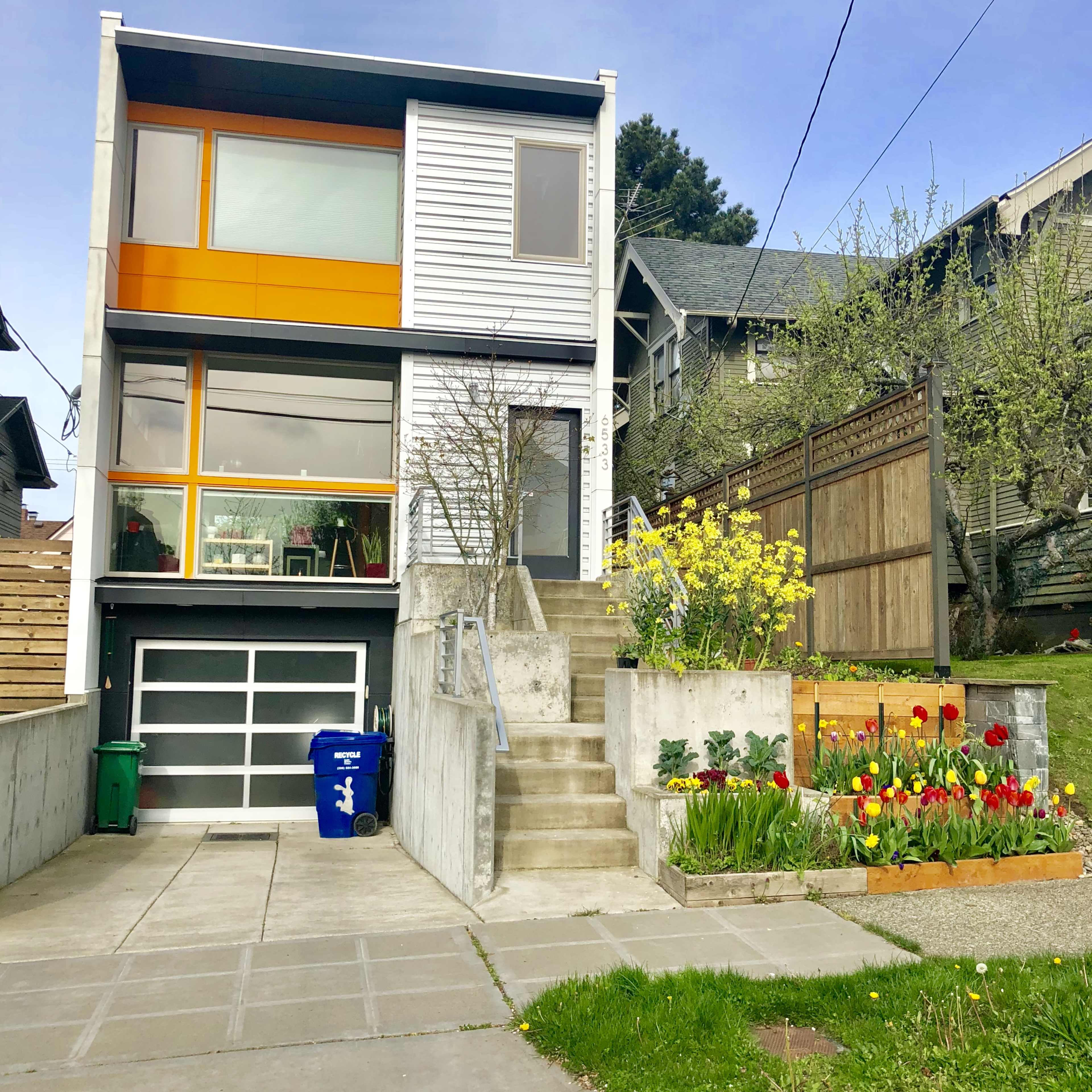 The image shows a modern two-story house with a mix of white and orange exterior, featuring a landscaped front yard with colorful flowers and a concrete pathway leading to the entrance.