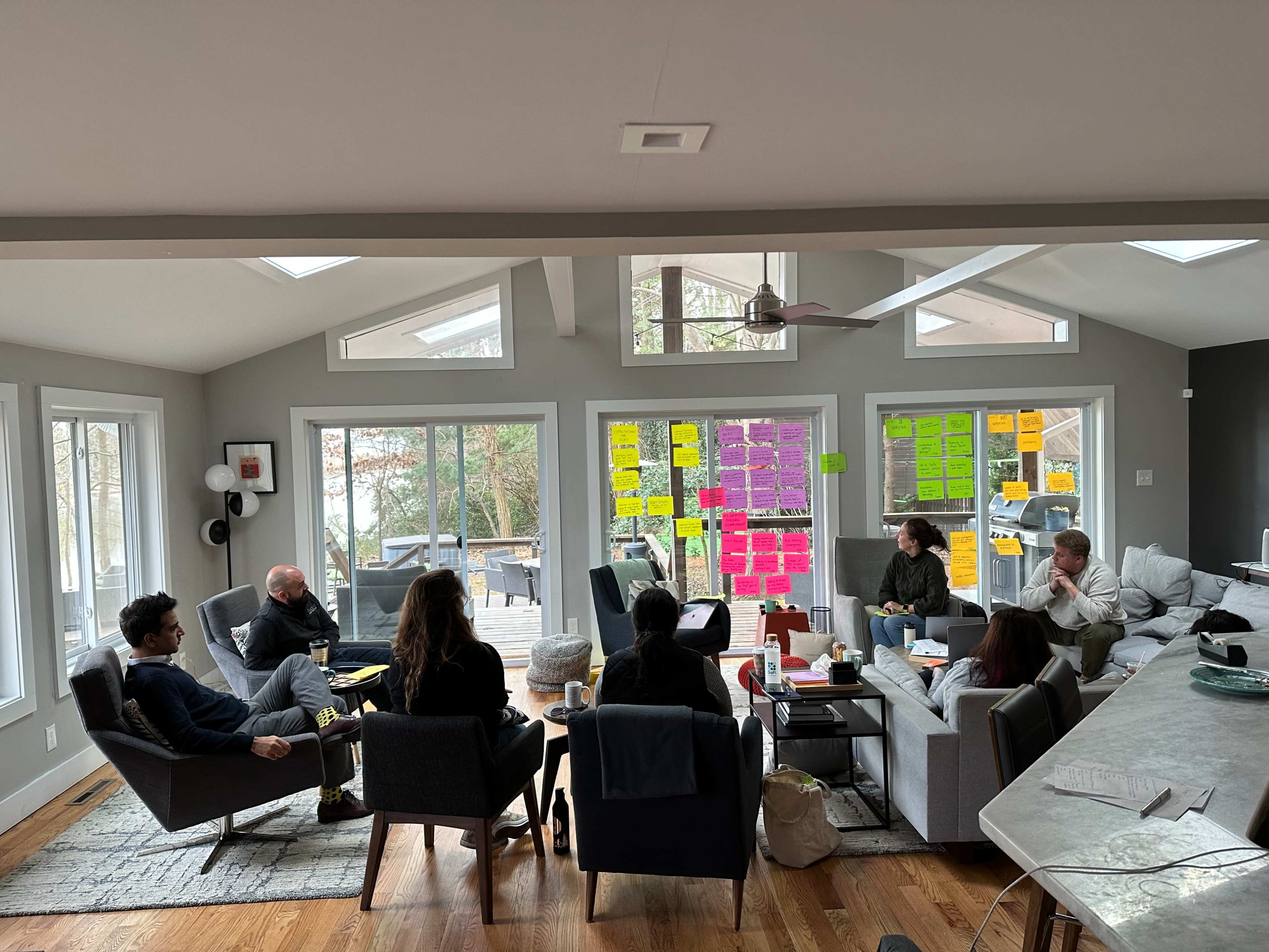 A group of people is seated in a spacious, well-lit room, discussing around a coffee table, with colorful sticky notes displayed on the windows.