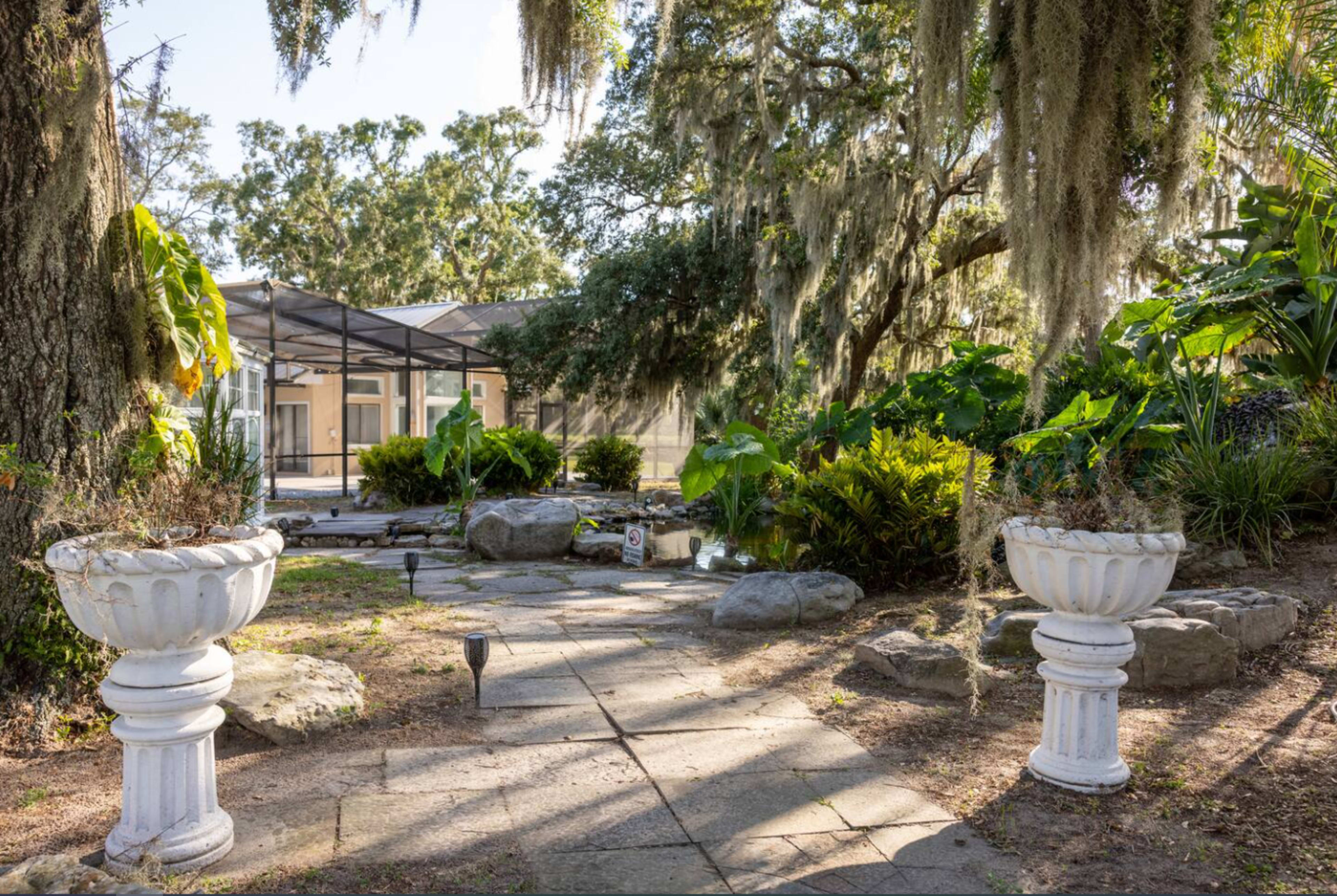 The image shows a garden path lined with white urns leading to a screened enclosure surrounded by lush greenery and trees with moss.
